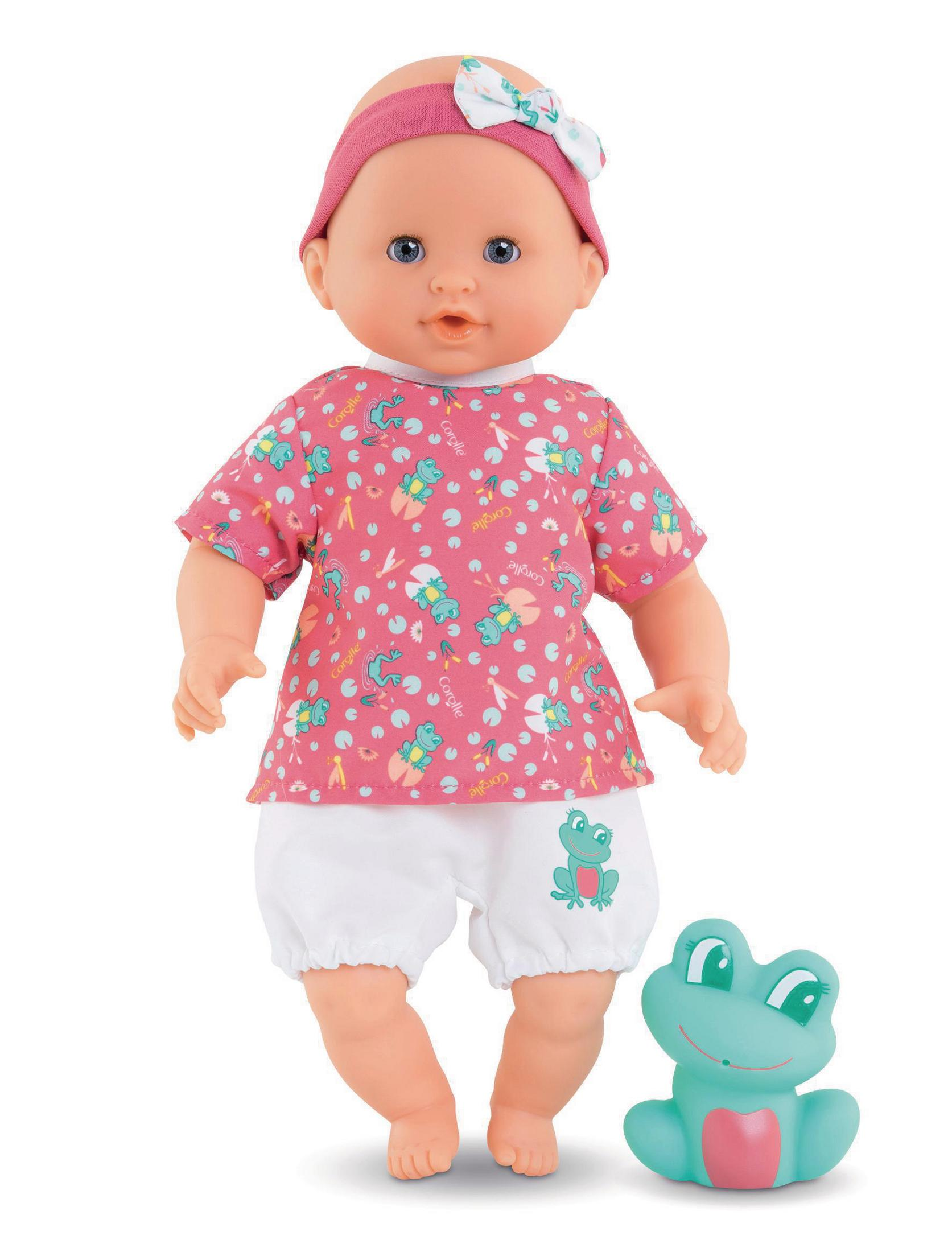 Eine Babypuppe mit einem pinken Hemd mit Frosch- und Seerosenmuster, weißen Shorts, einem pinken Stirnband und einem grünen Froschspielzeug.