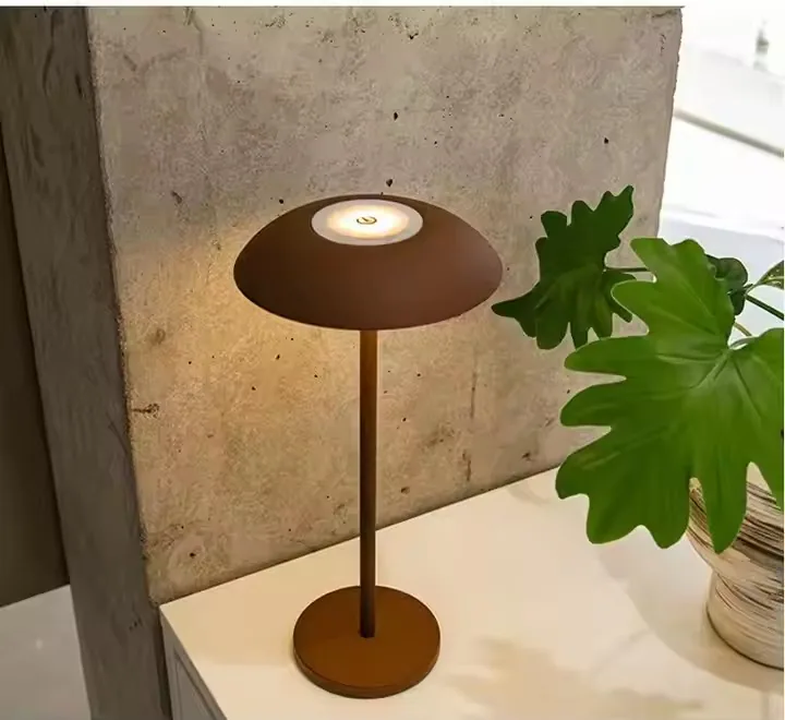 Braune Tischlampe mit rundem Lampenschirm und Sockel auf weißem Tisch, daneben eine Pflanze.