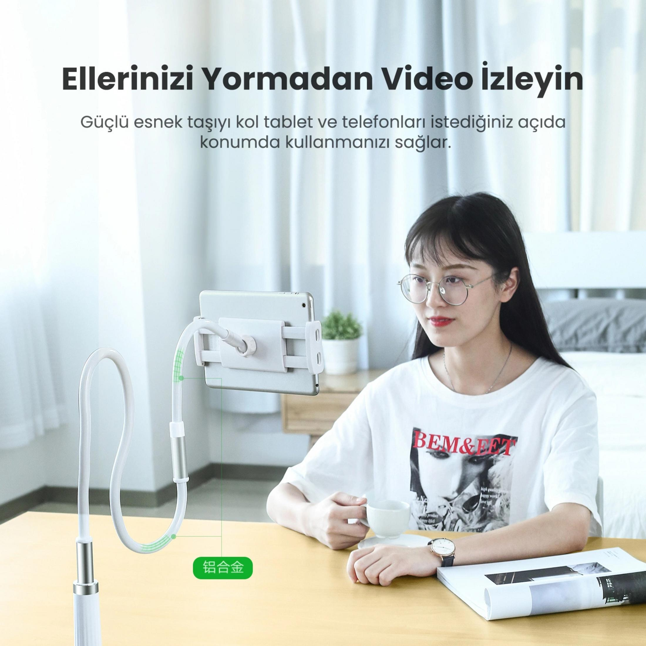 Kobieta w okularach siedzi przy stole, trzymając filiżankę. Obok niej stoi stojak na tablet.