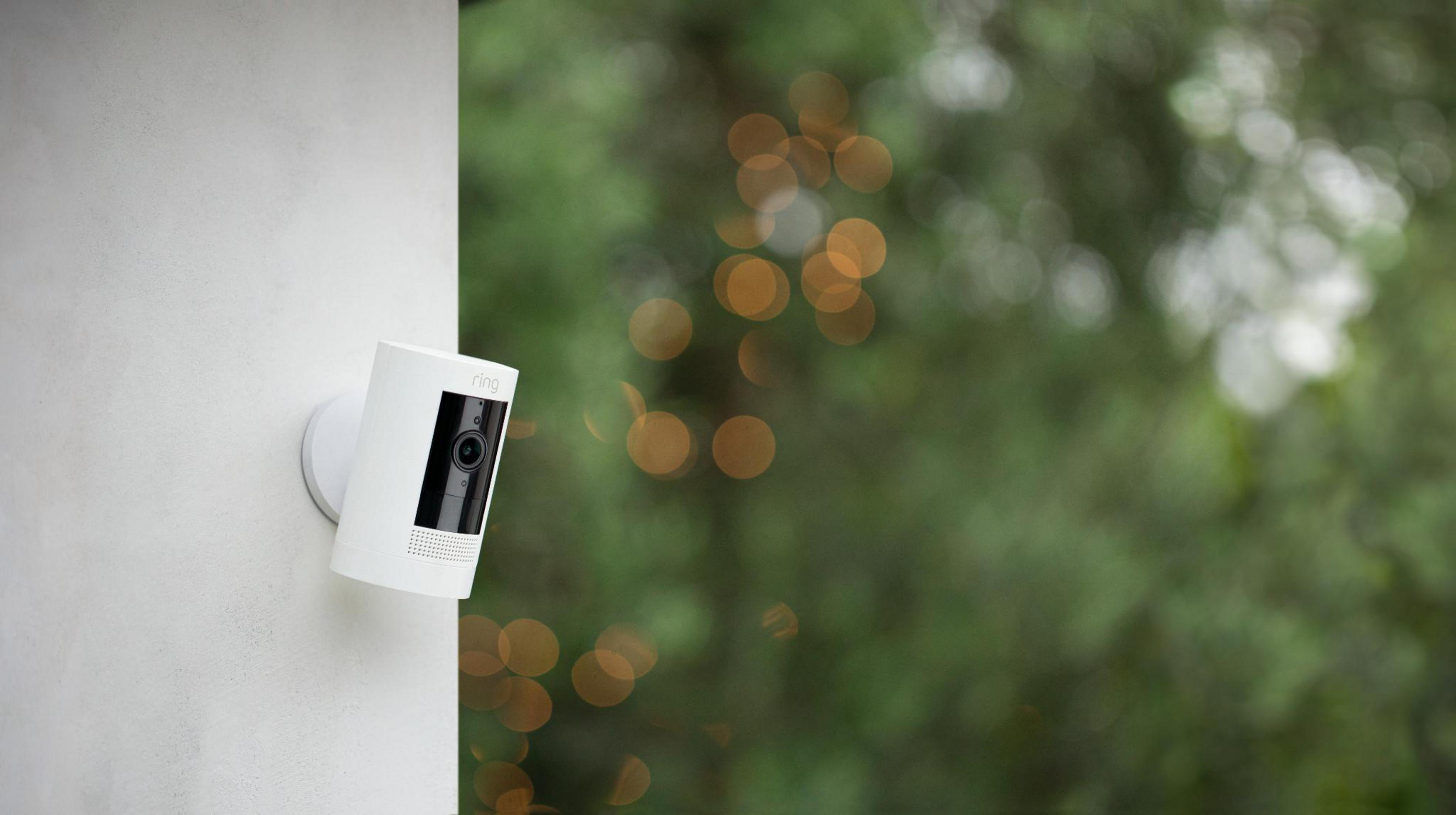 Telecamera di sicurezza Ring bianca montata su muro bianco, sfondo verde sfocato.