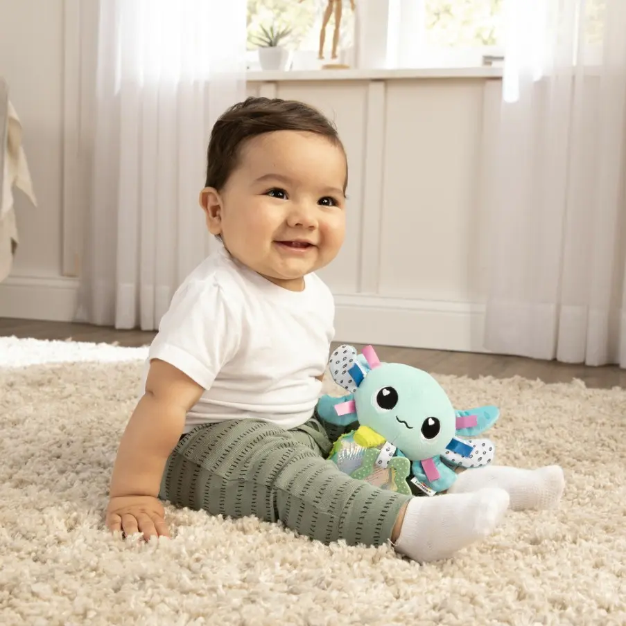 Un bebé se sienta en una alfombra junto a un peluche.