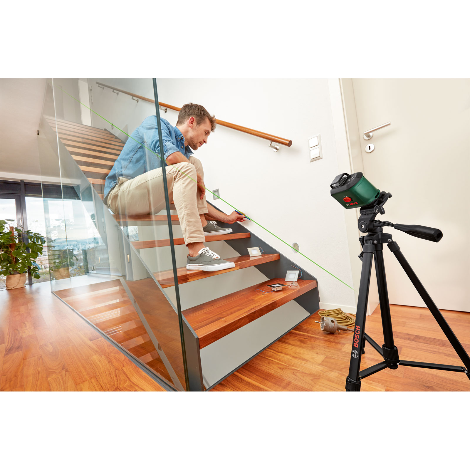 Mann auf Holztreppe mit Glasgeländer, der mit Laserpegelmarkierungen arbeitet.