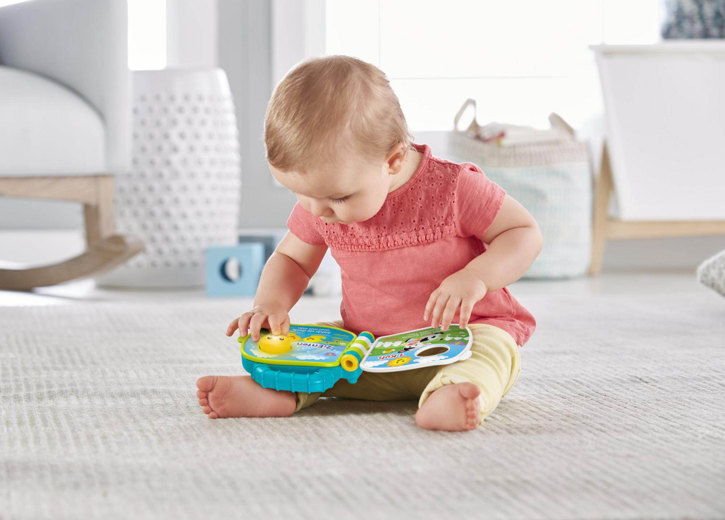 Ein Baby in einem rosa Hemd spielt mit einem bunten Spielzeugbuch auf dem Teppich.