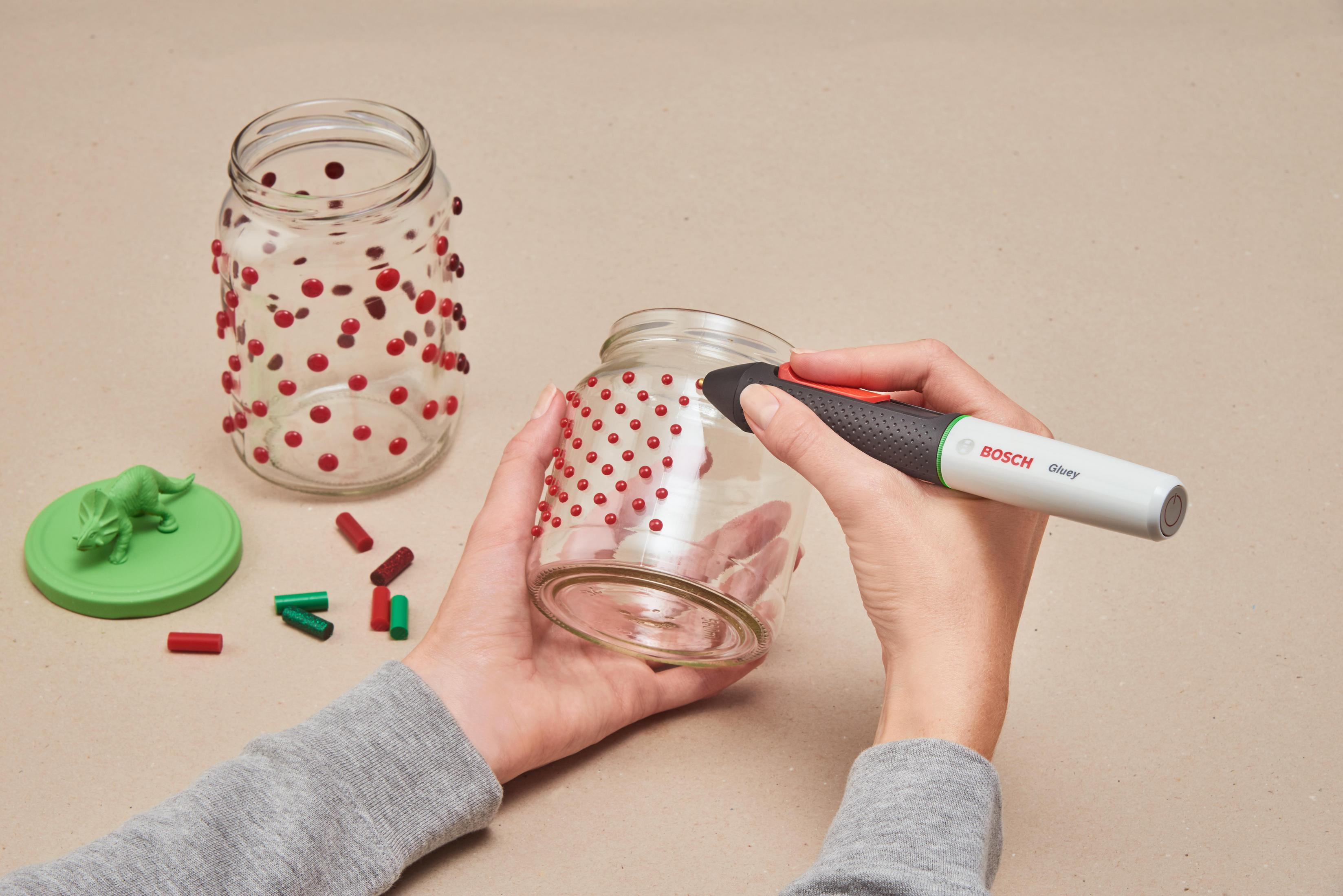 Eine Hand benutzt einen Bosch Gluey Klebestift, um rote Punkte auf ein Glas aufzutragen.