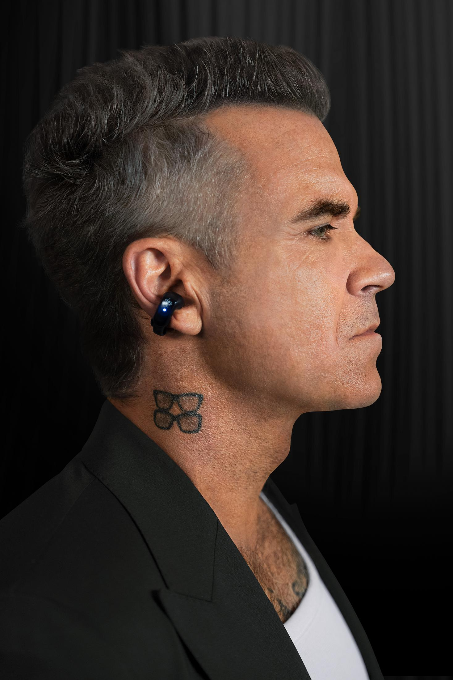 Hombre con cabello gris viste traje negro, audífono azul y tatuaje de gafas.