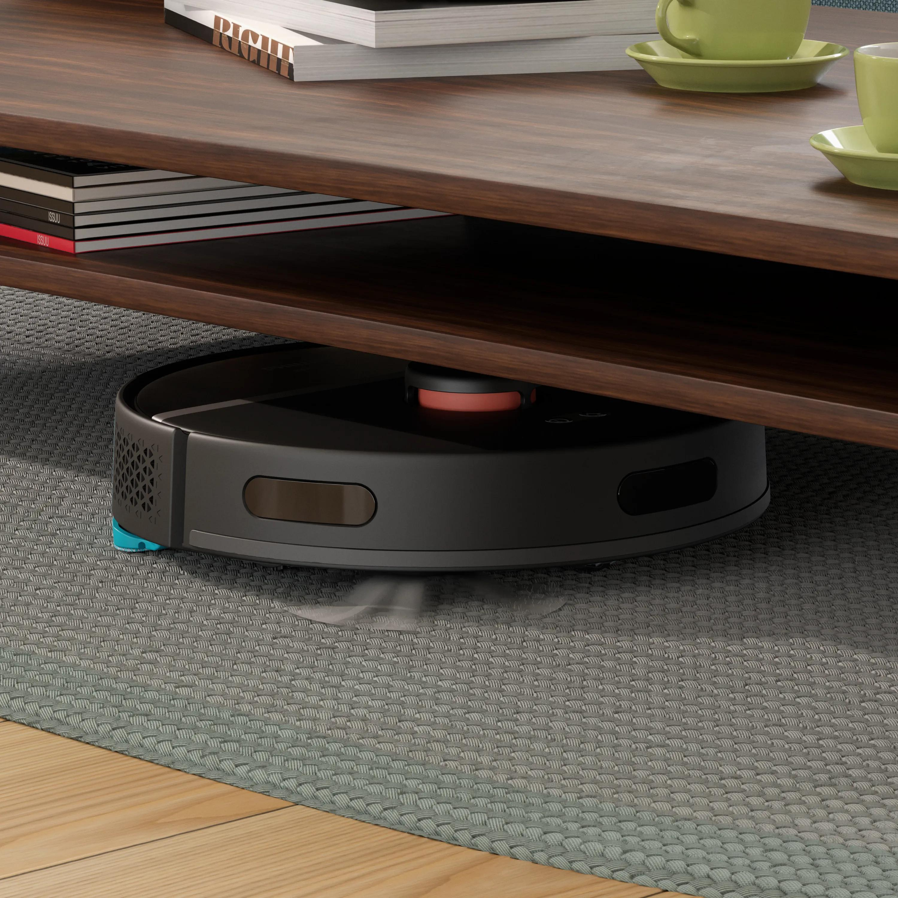 Siyah bir robot süpürge, kitaplar ve yeşil fincanların olduğu ahşap masanın altında temizlik yapıyor.