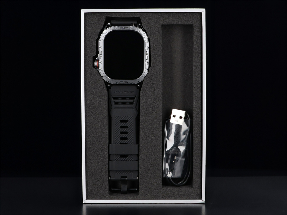 Czarny smartwatch z kablem ładującym, w białym pudełku.