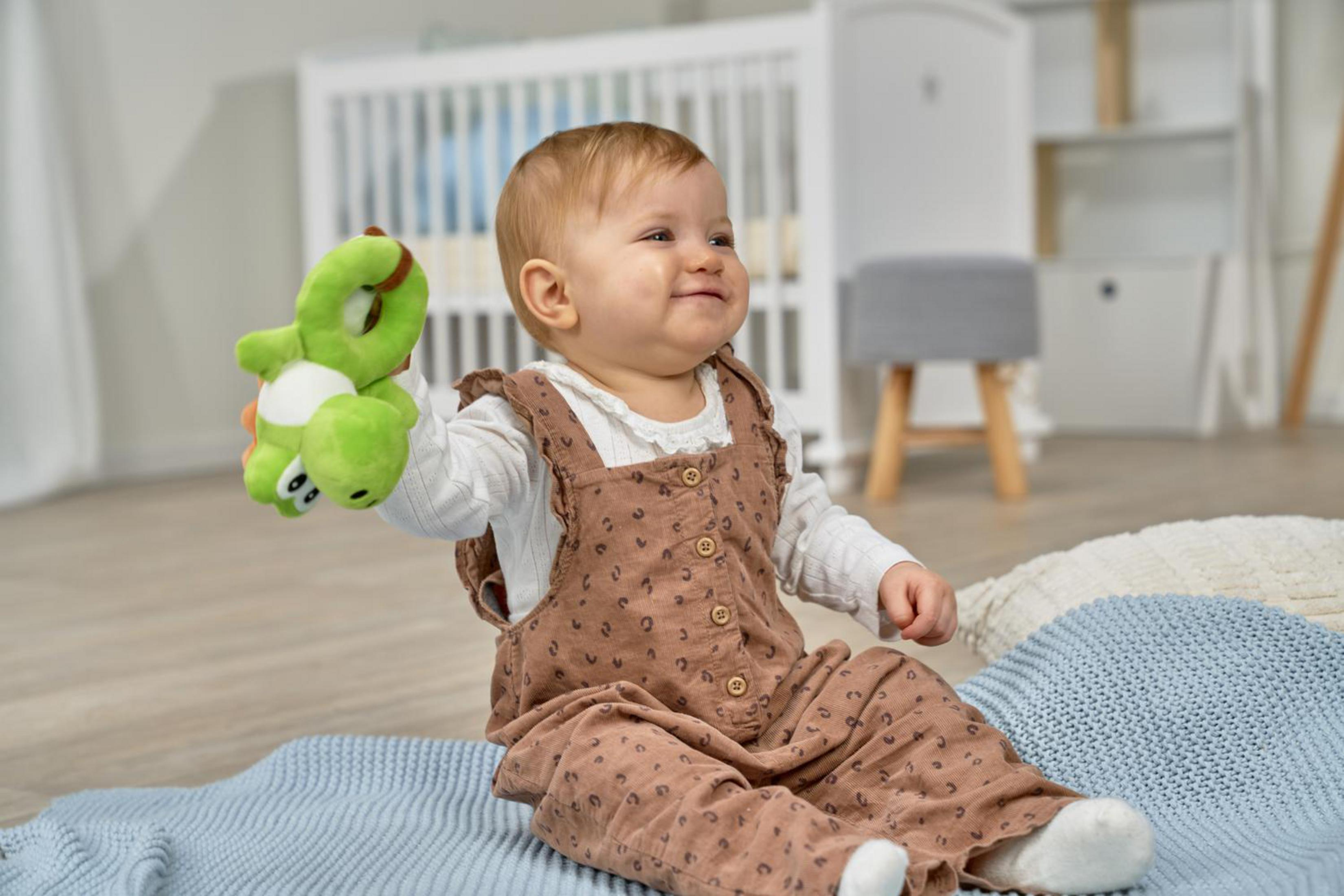 Ein Baby in Latzhosen hält ein grünes Spielzeug, sitzt auf einer Decke.