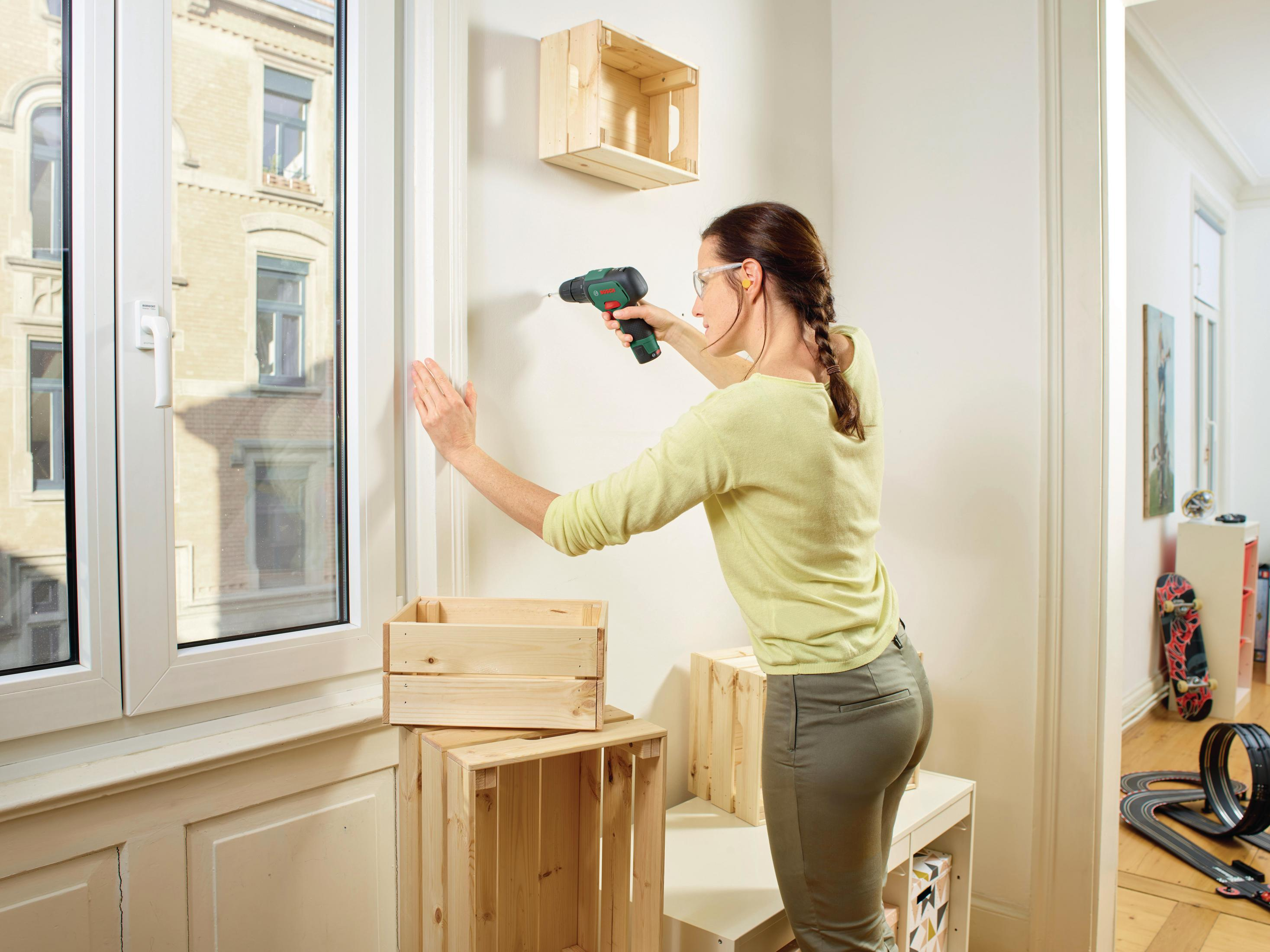 Frau bohrt Holzregale an Wand. Fenster, Kisten, Skateboard, Autorennbahn im Raum.