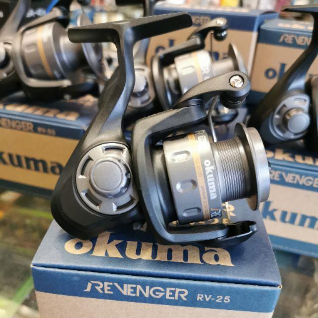 Siyah Okuma Revenger RV-25 olta makarası mavi kutusunun üzerinde duruyor. Arkada başka makaralar var.