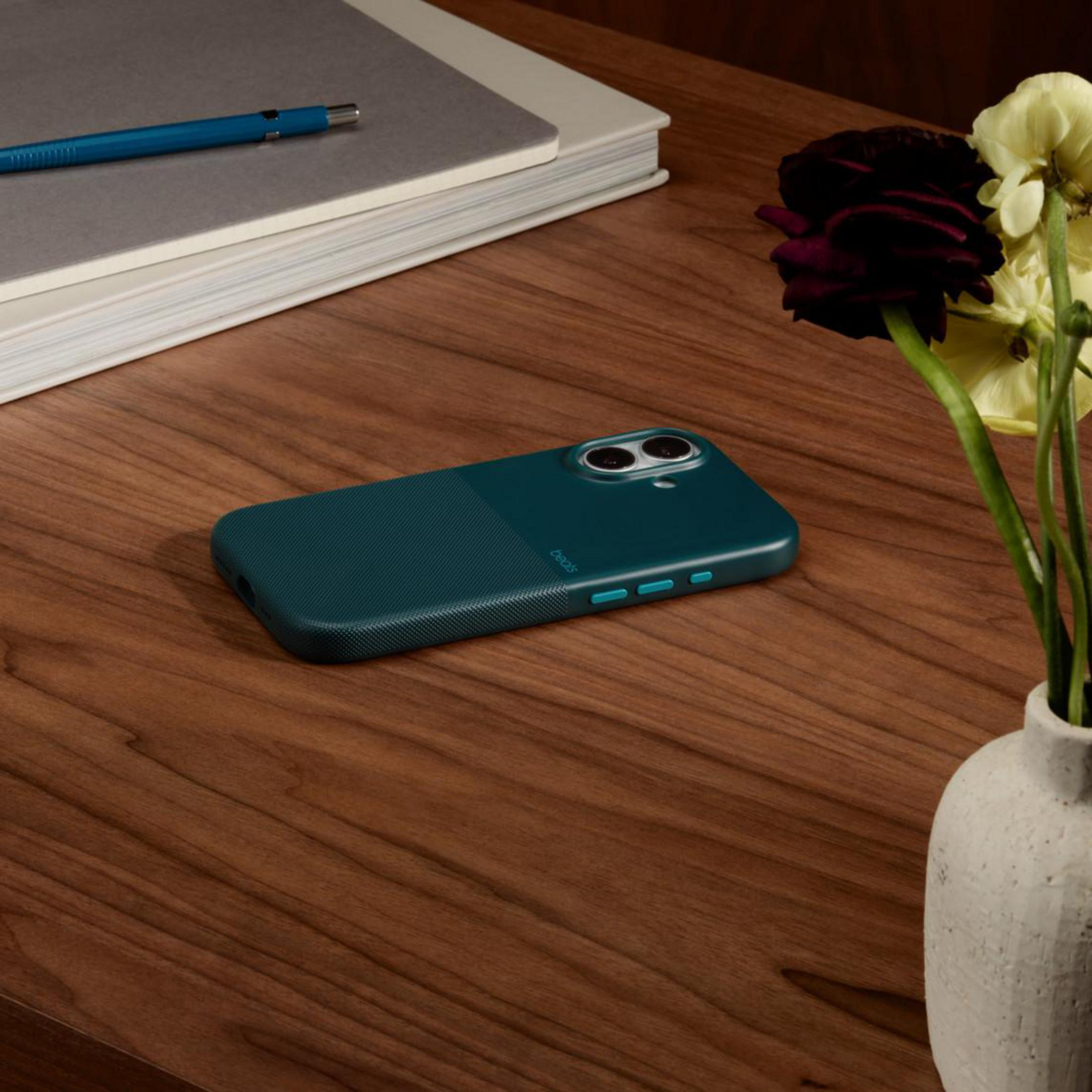 Una custodia per telefono verde acqua scuro su una scrivania di legno accanto a fiori e un taccuino.