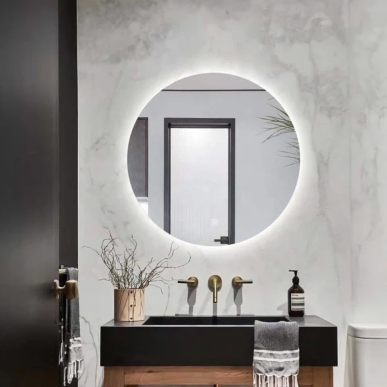 Een modern wastafelmeubel met zwart bassin, gouden kranen en een verlichte ronde spiegel.