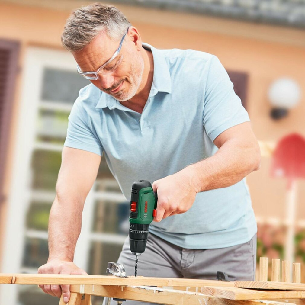 Mann mit Schutzbrille bohrt Holz mit grüner Bohrmaschine im Freien.