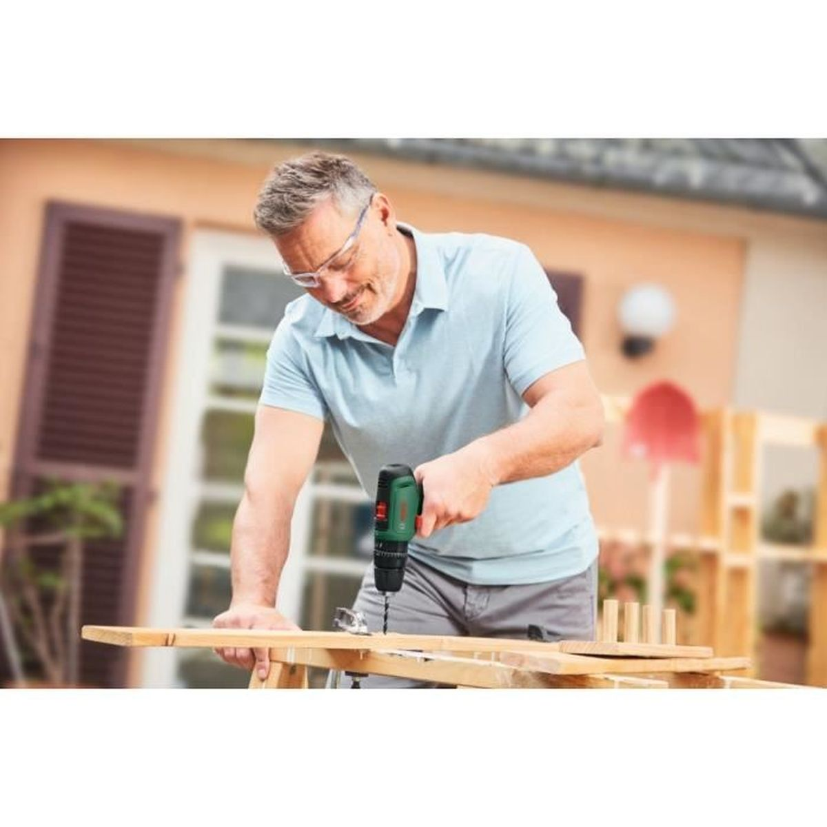 Mann bohrt in Holz. Er trägt eine blaue Poloshirt und Schutzbrille.