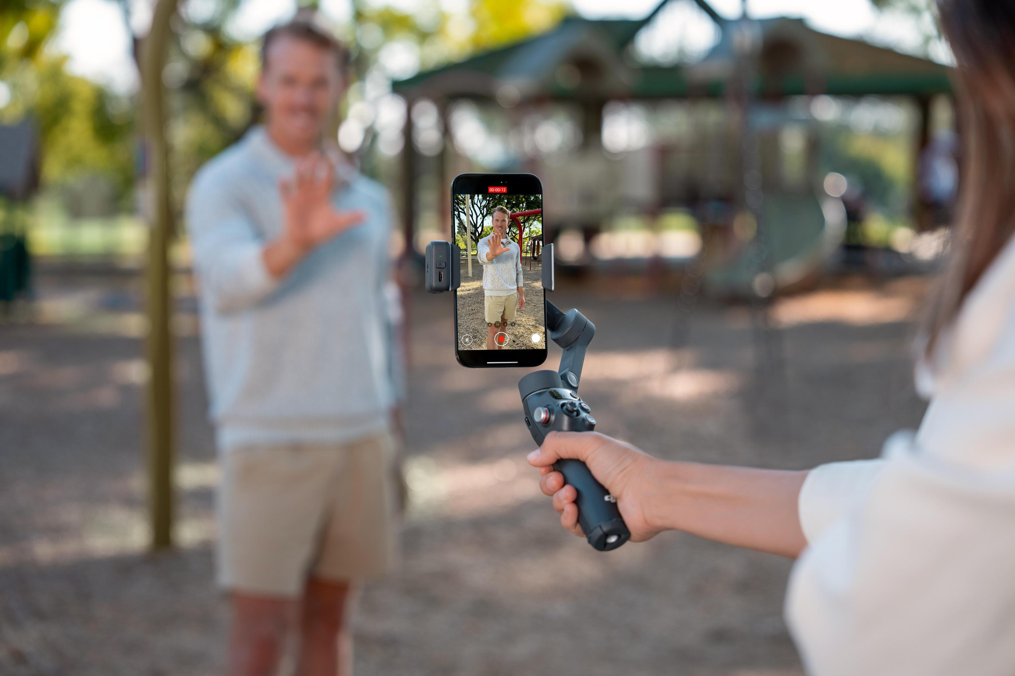 Mano tiene gimbal con telefono, uomo saluta sullo sfondo.