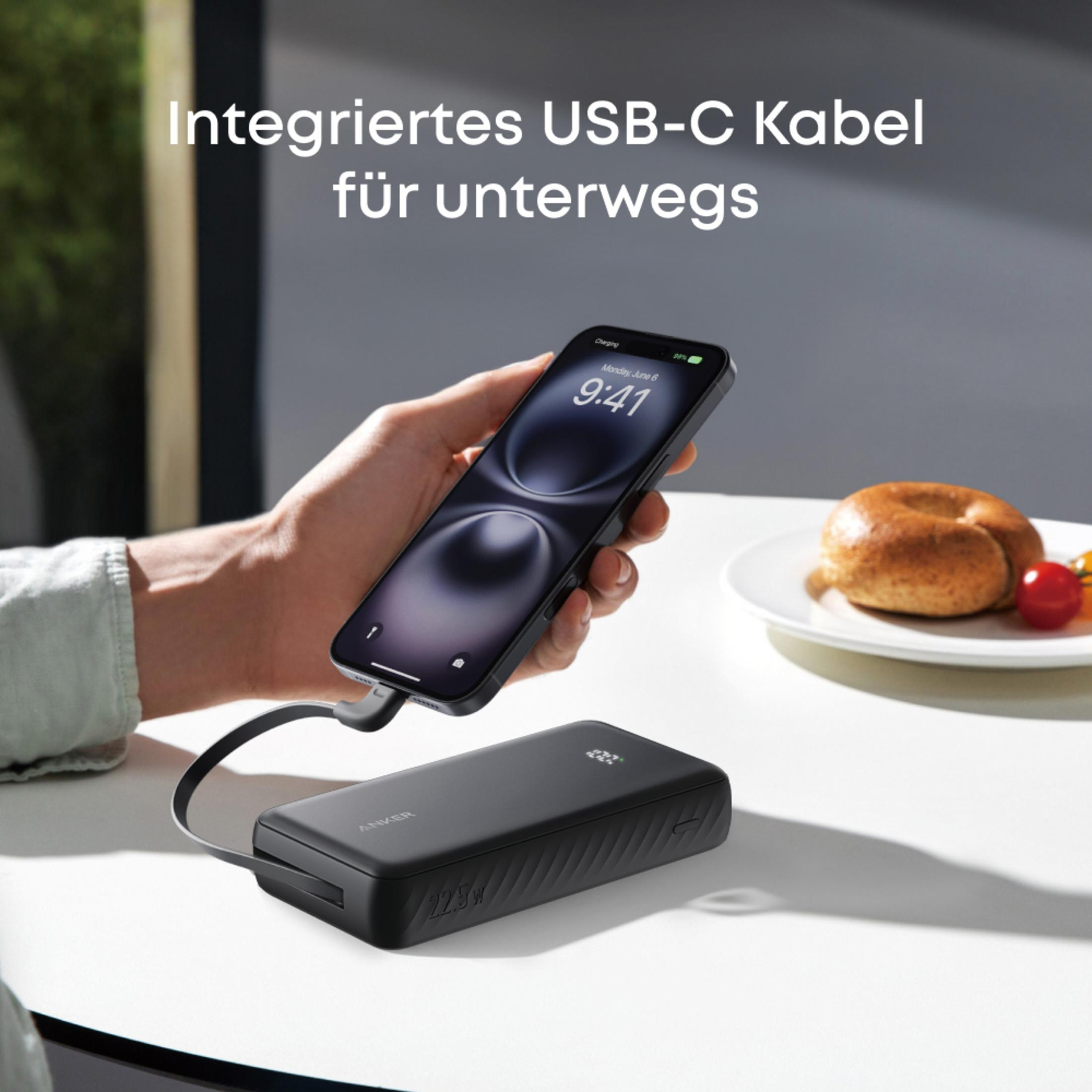 Power bank Anker negra carga un iPhone sobre una mesa con bagel y tomates.