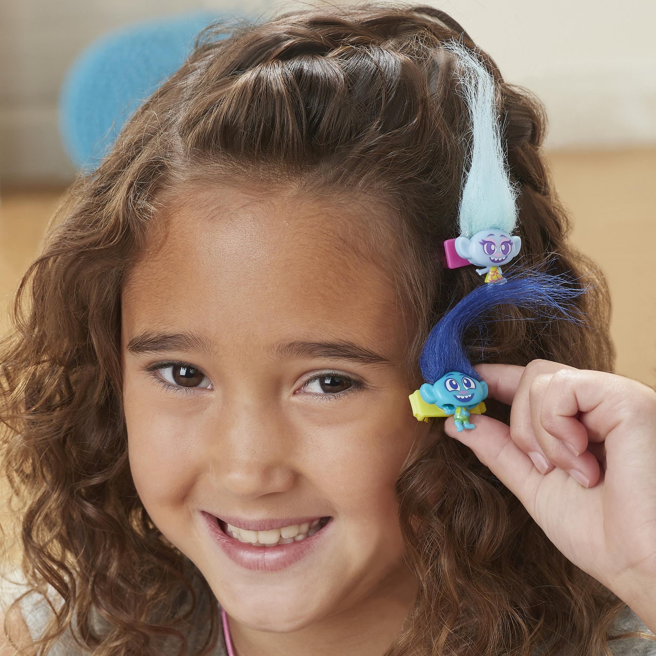 Une fille aux cheveux bruns sourit, avec deux figurines Trolls dans ses cheveux.