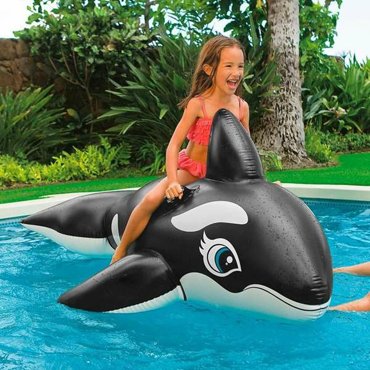 Una bambina cavalca un'orca gonfiabile in piscina, circondata dal verde.
