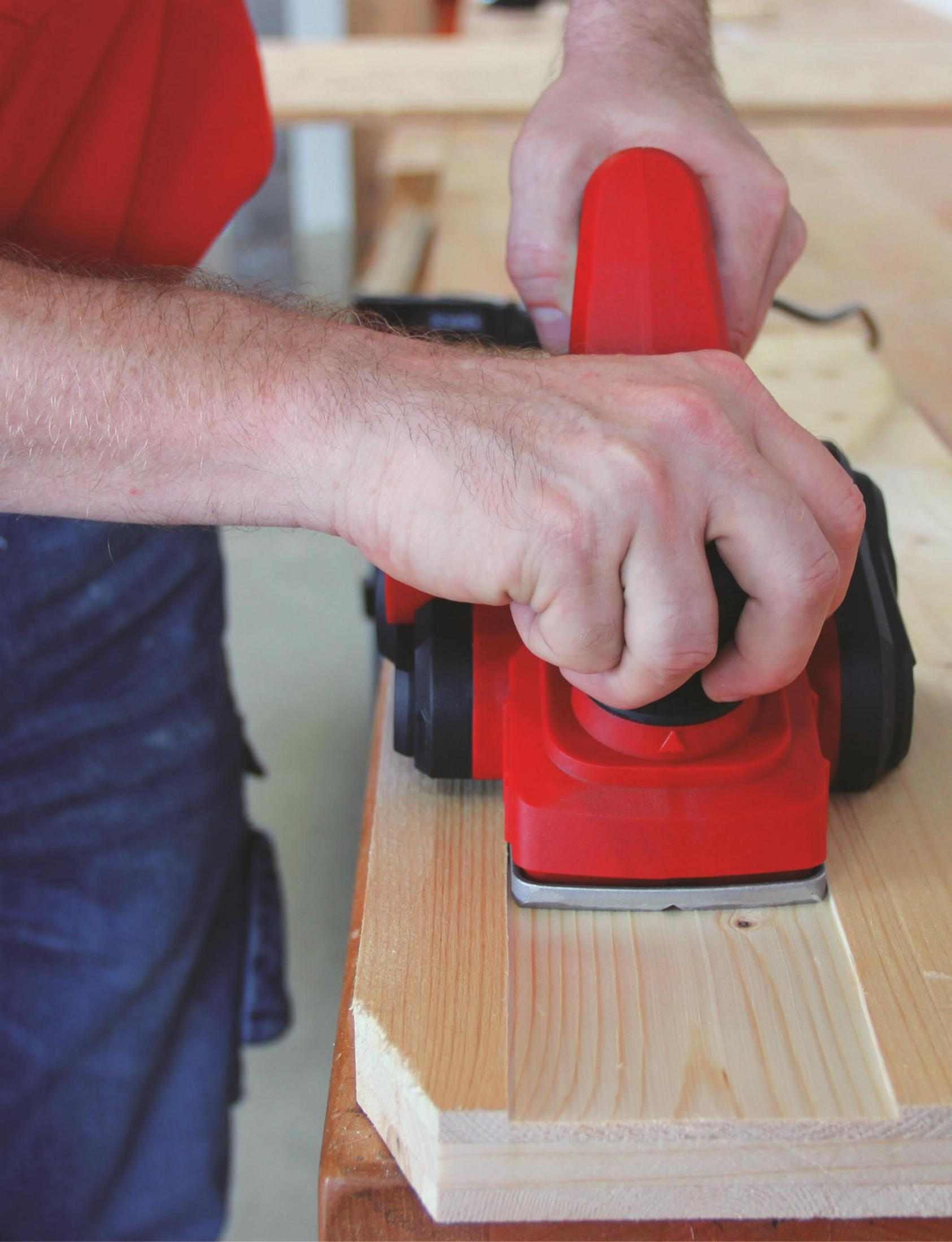 Un uomo pialla il legno con una pialla rossa su un banco da lavoro.