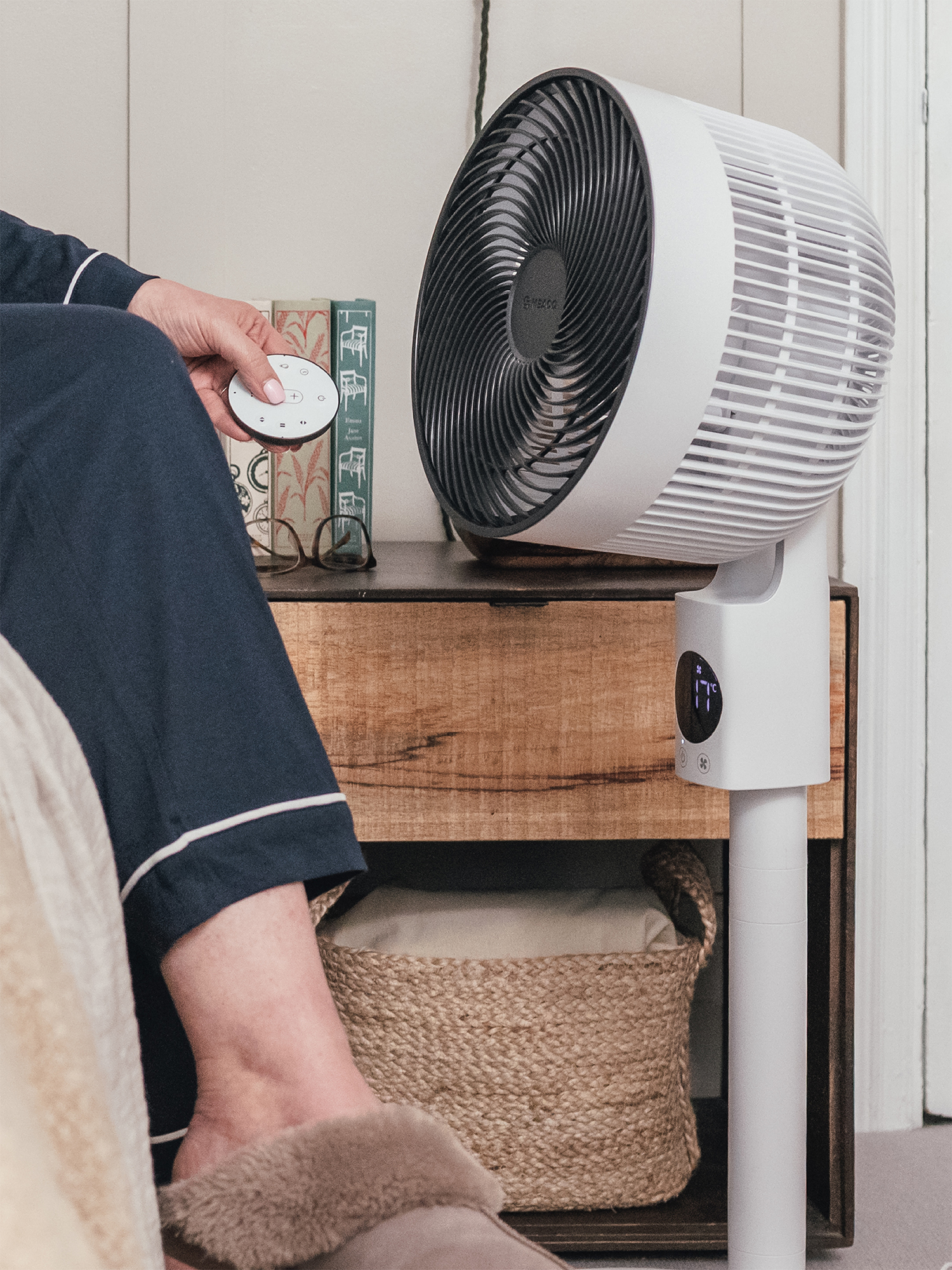 Una persona sostiene un mando a distancia junto a un ventilador en una mesita de noche.