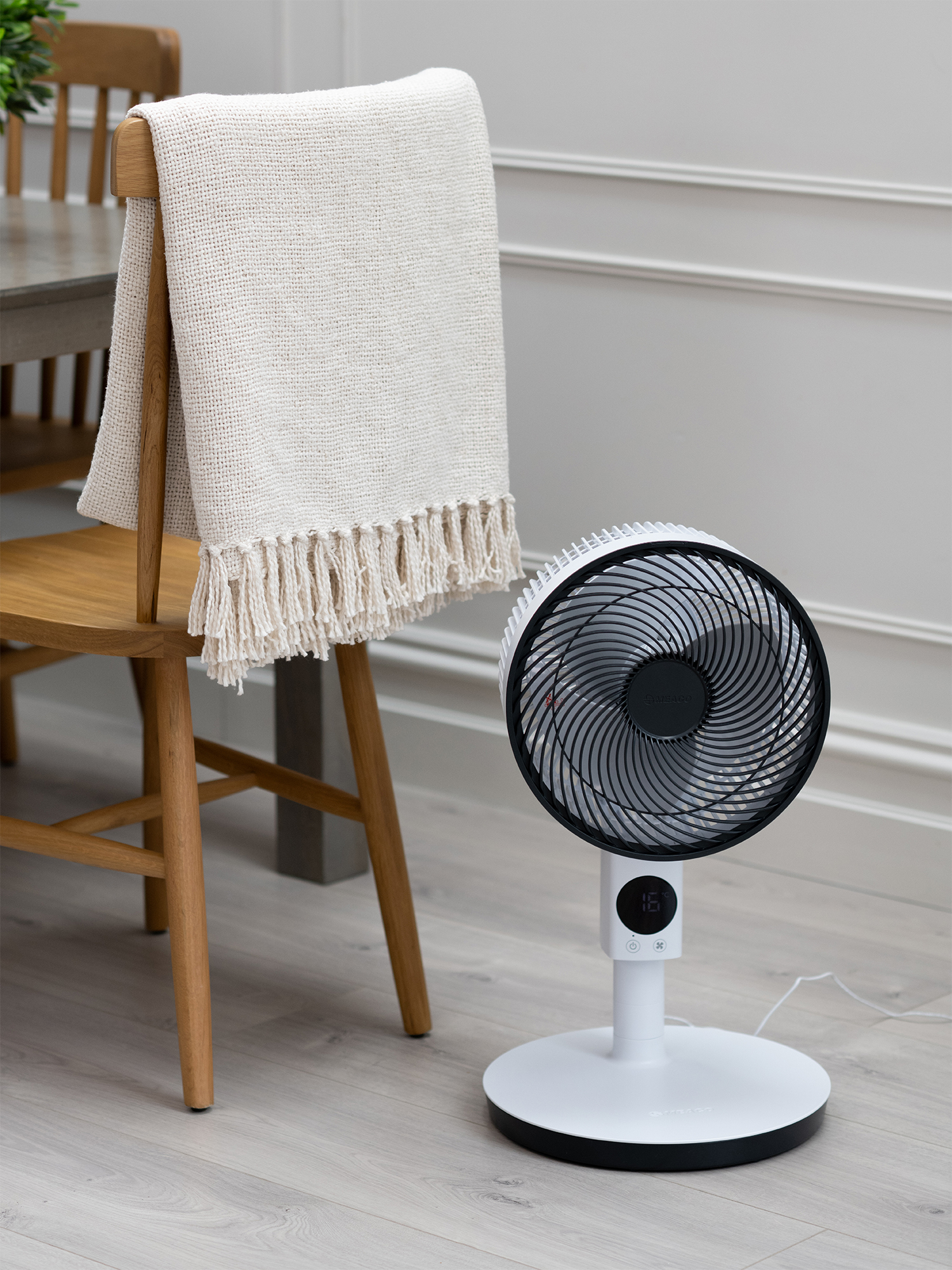 Un ventilador blanco está sobre un suelo de madera junto a una silla de madera con una manta.