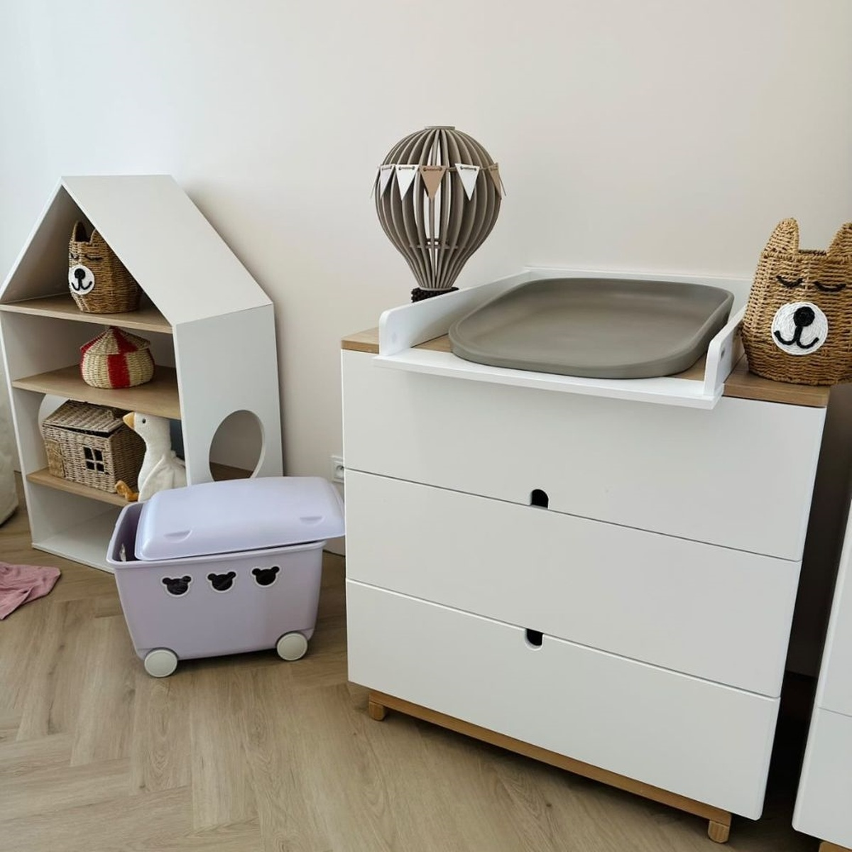 Ein Kinderzimmer mit einem Wickeltisch, einem Bücherregal in Hausform und einer Spielzeugkiste.