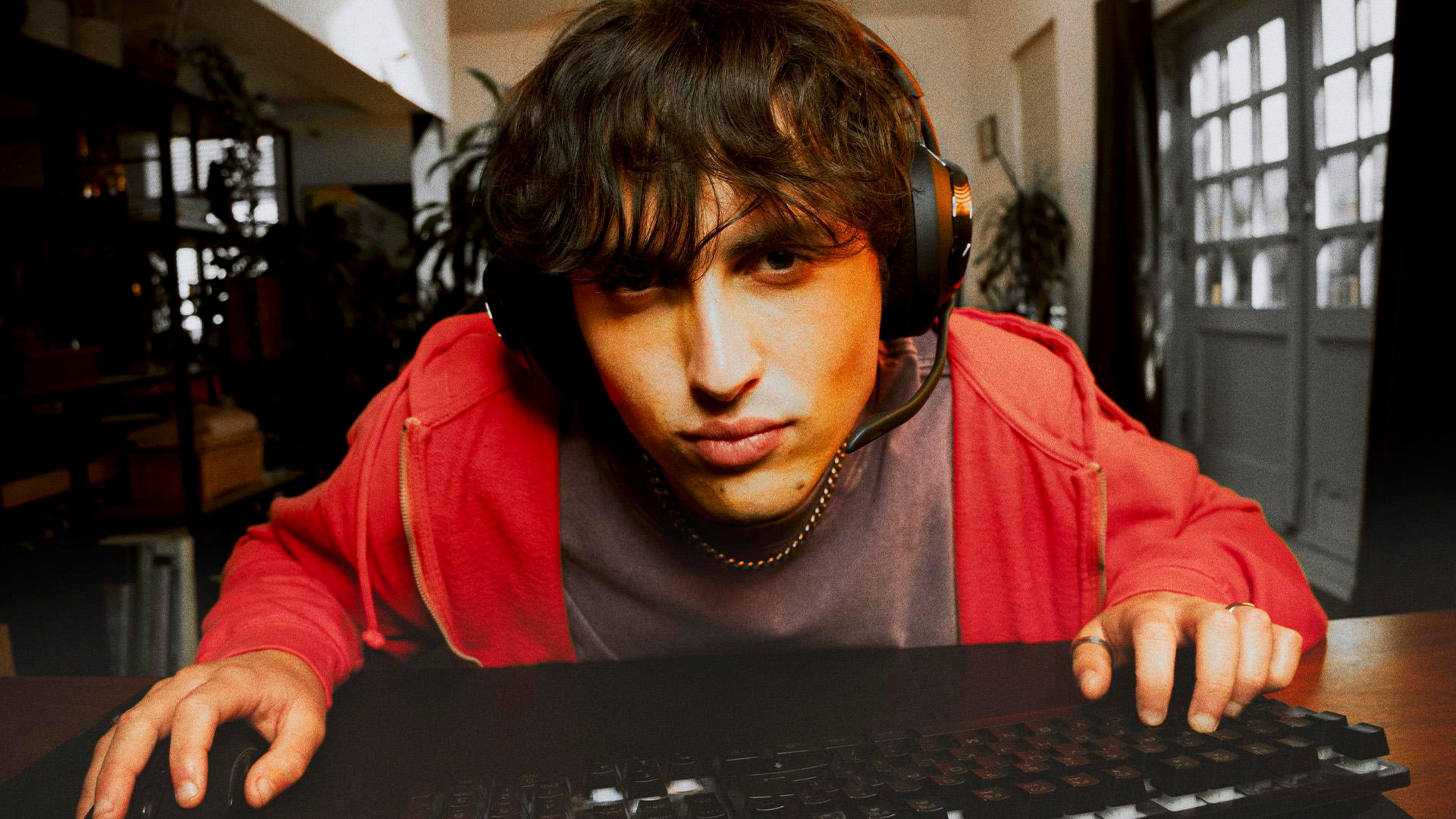 Un giovane con cuffie gioca al computer. Indossa una felpa rossa.