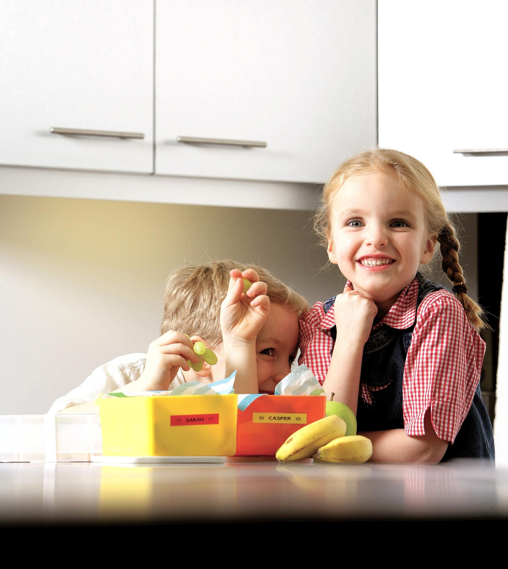 Zwei Kinder stehen an einer Küchentheke mit Lunchboxen, Obst und Snacks.