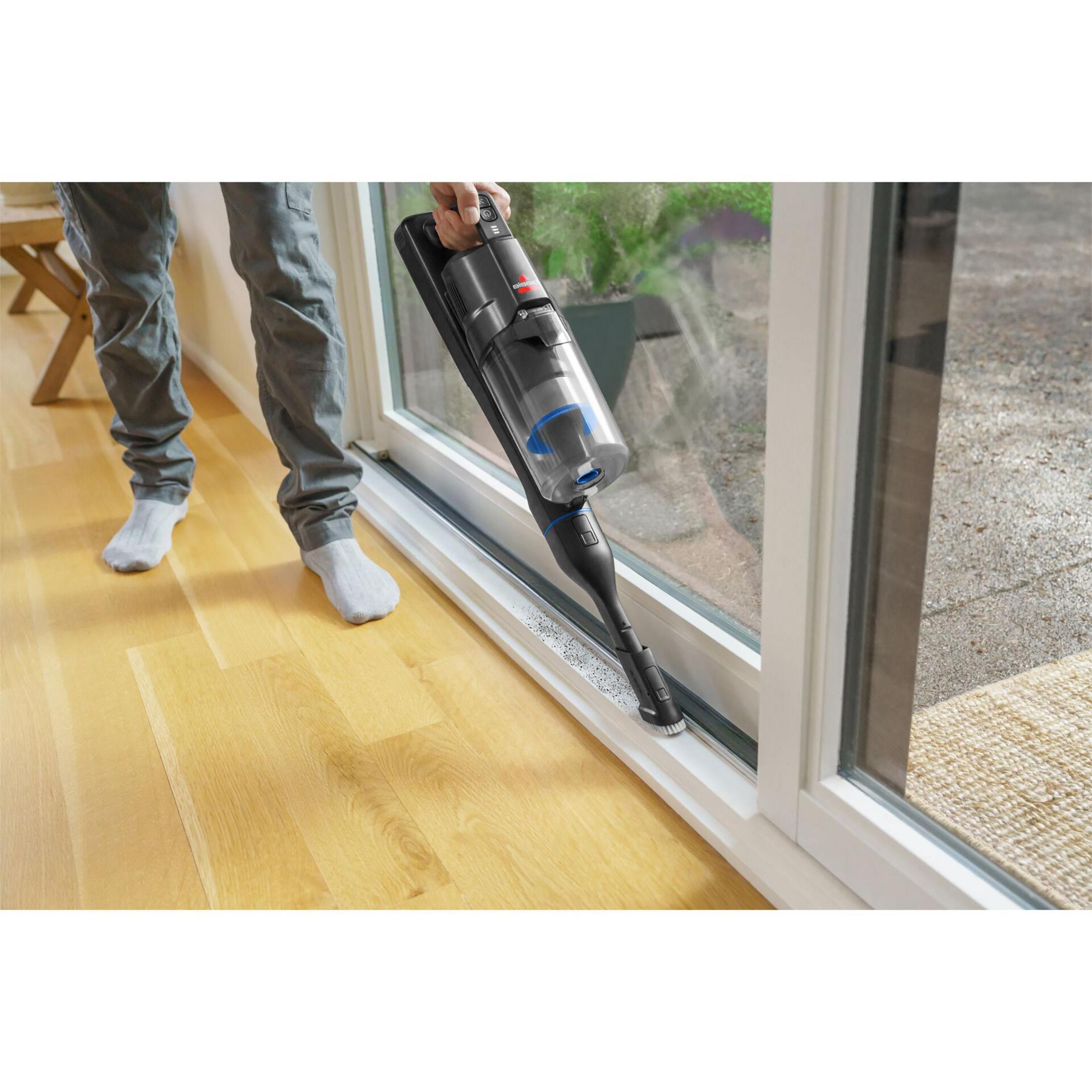 Person reinigt Fensterrahmen mit einem schwarzen Handstaubsauger auf Holzboden.