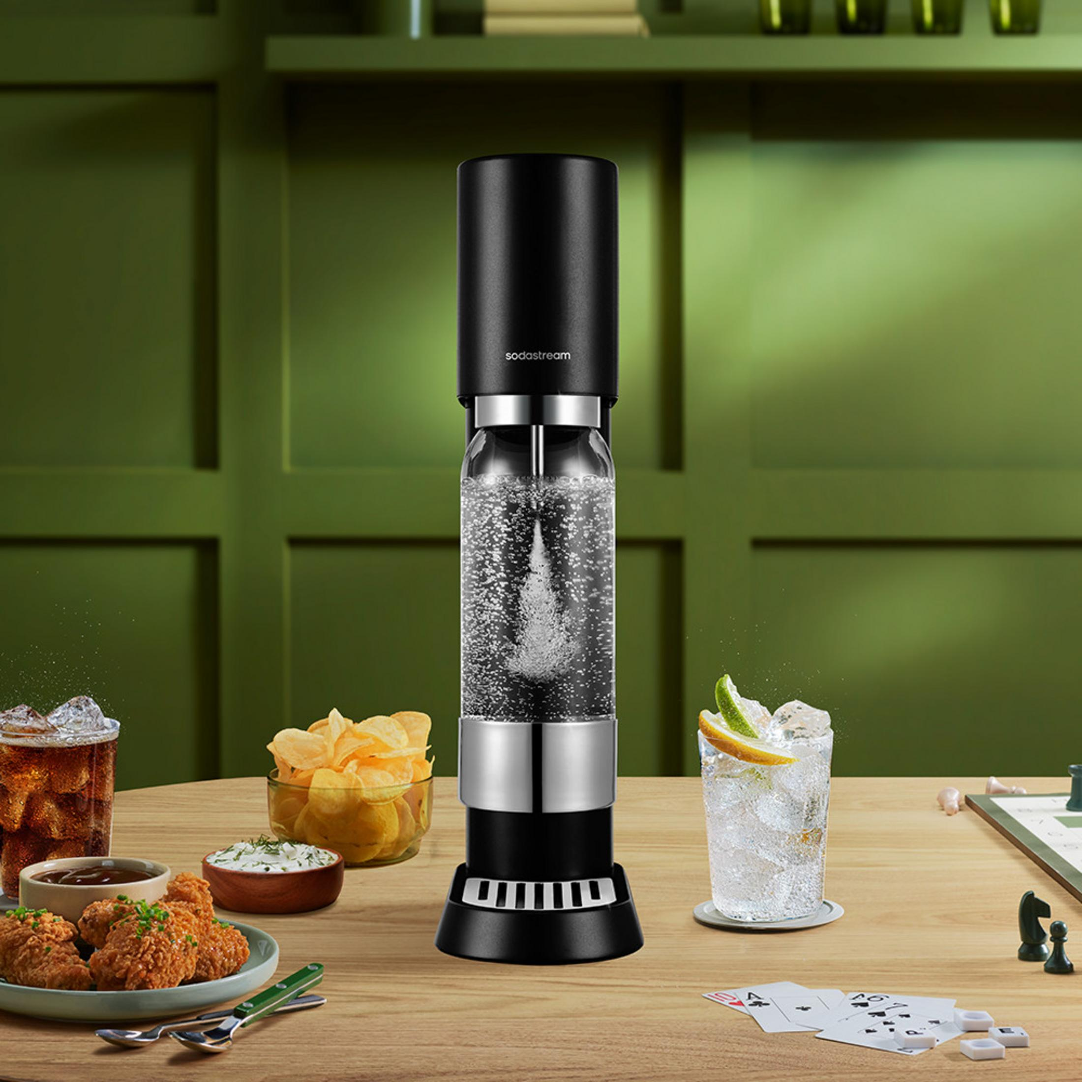 Ein schwarzer SodaStream mit sprudelndem Wasser steht auf einem Holztisch mit Snacks und Getränken.