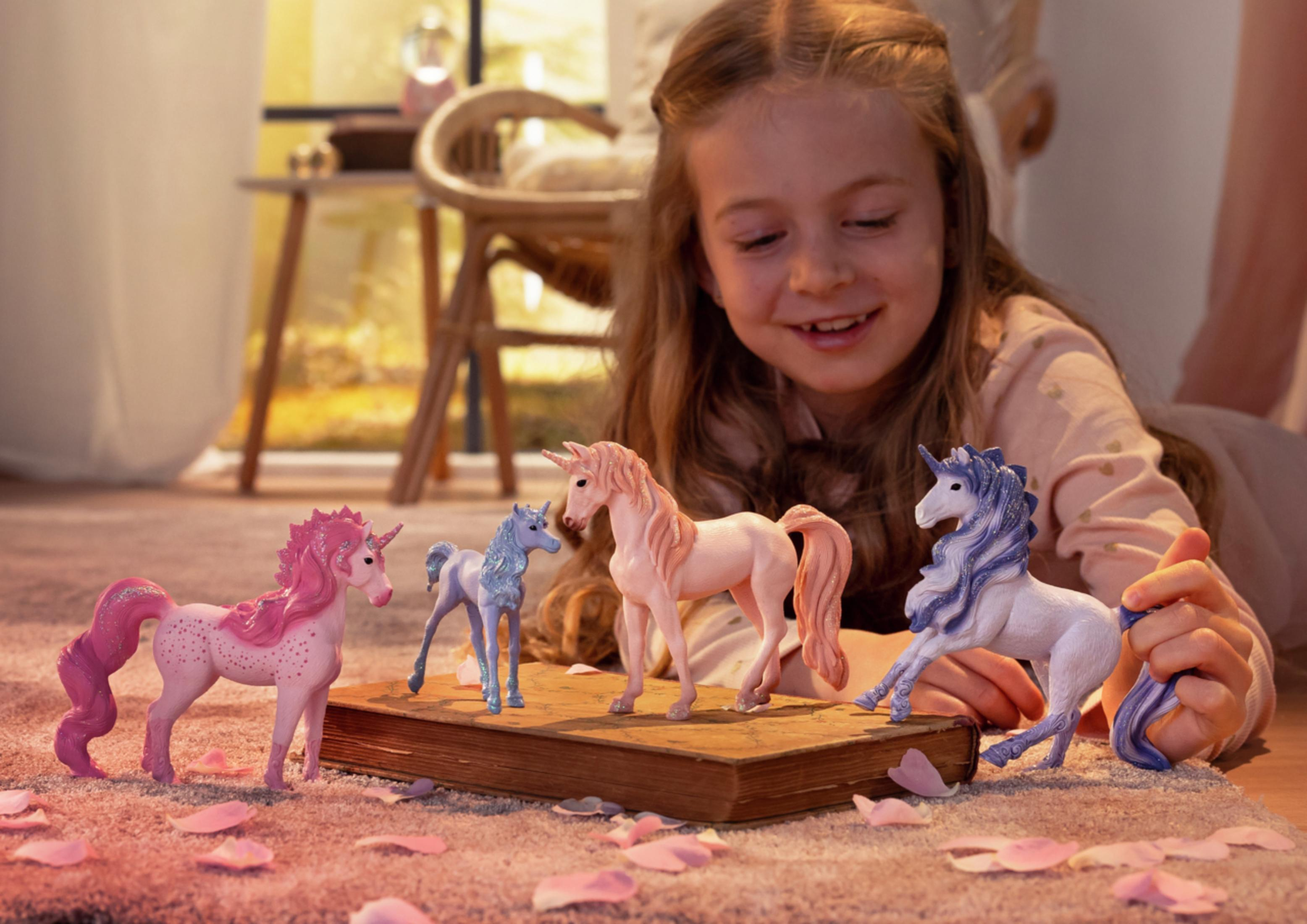 Ein Mädchen spielt mit vier Einhorn-Figuren auf einem Teppich mit Blütenblättern.