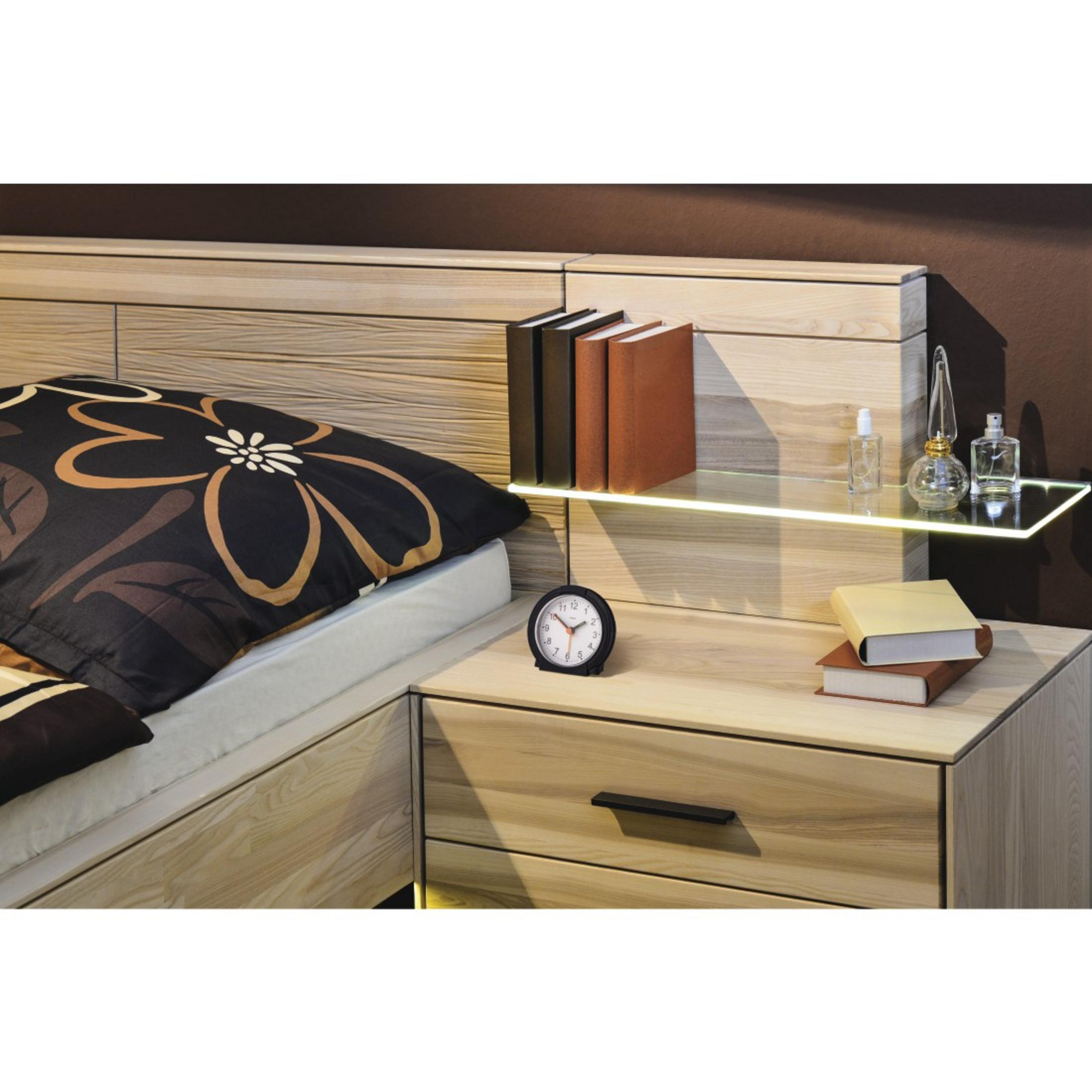 Un comodino in legno chiaro con cassetti, una mensola con libri e boccette di profumo.