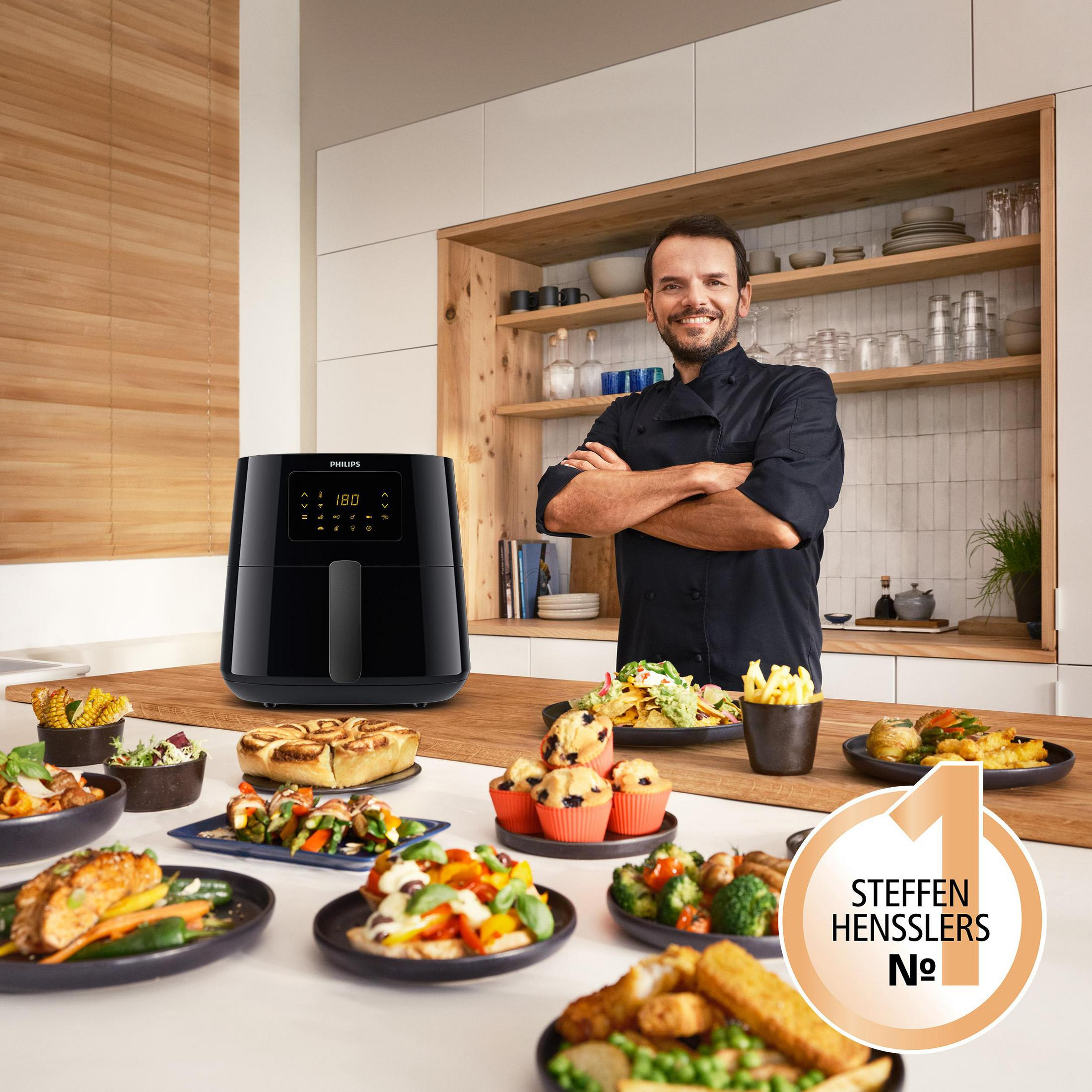 Uno chef si trova in una cucina accanto a una friggitrice ad aria nera e molti piatti.