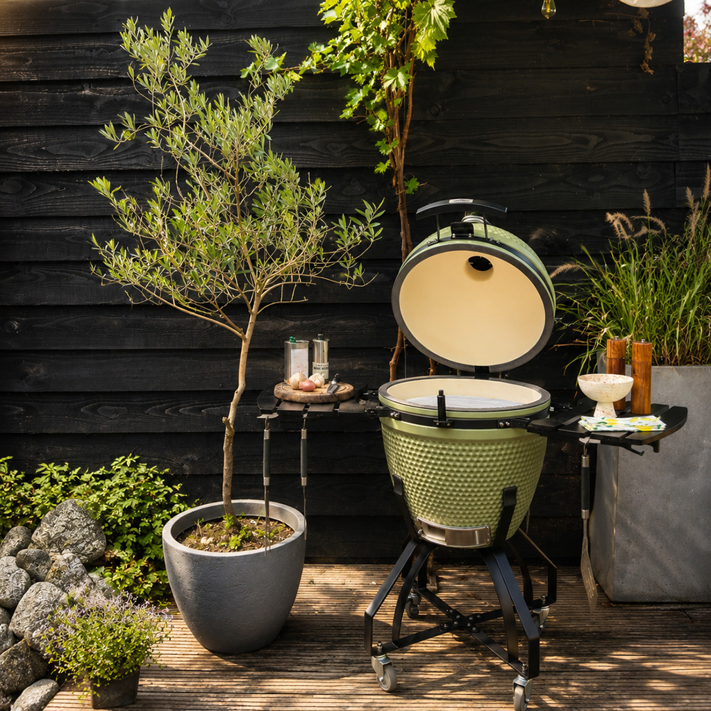 A green ceramic grill sits on a wooden deck beside an olive tree.
