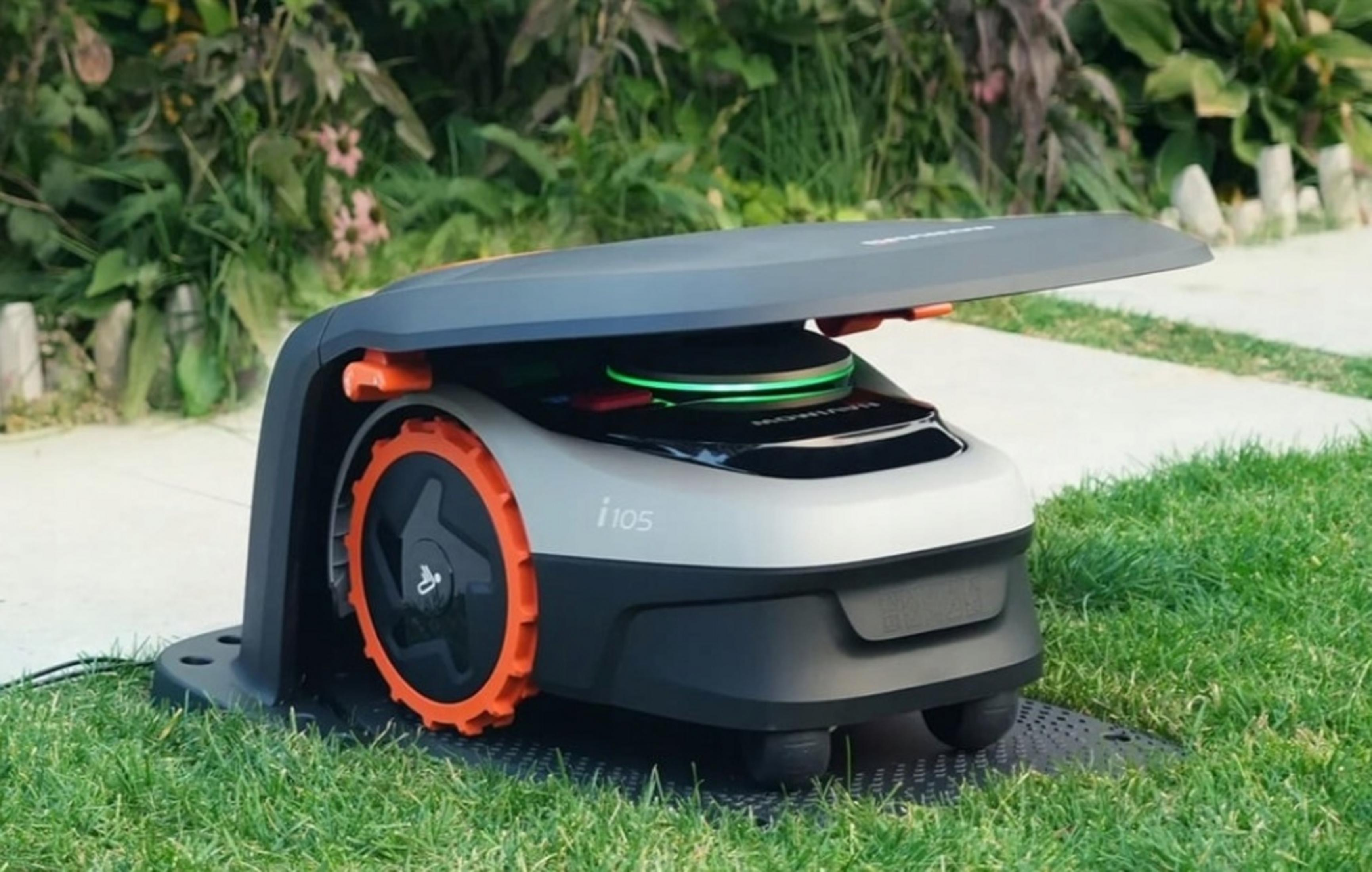 Ein moderner Mähroboter mit orangefarbenen Rädern steht auf einem Rasen vor Grünpflanzen.