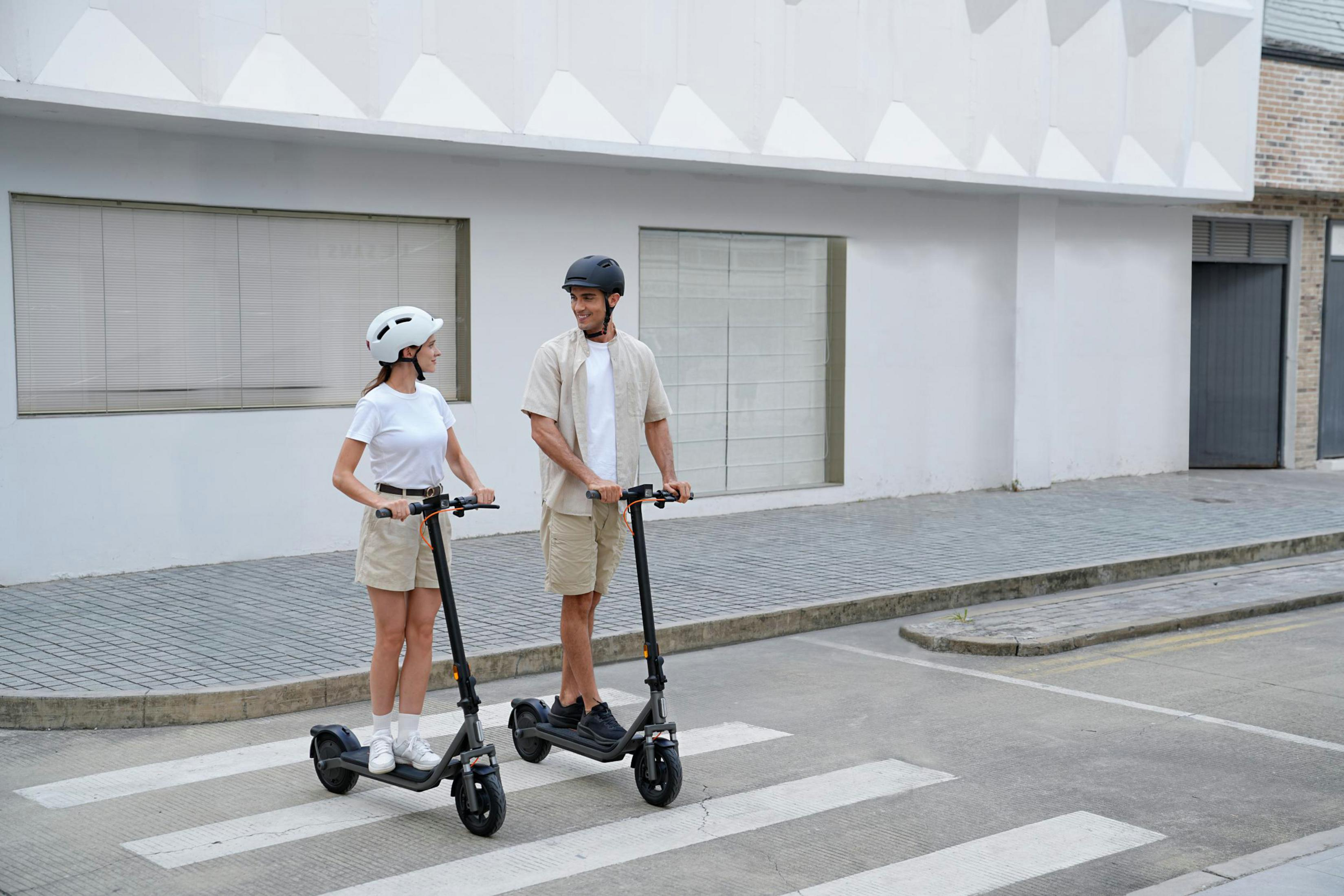 Uomo e donna su e-scooter su strisce pedonali di fronte a edificio.