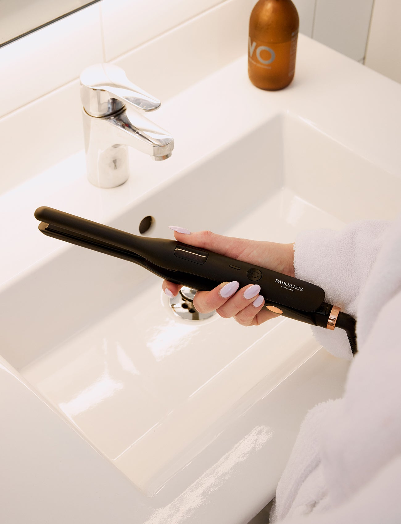 Una mano sostiene un alisador de pelo negro sobre un lavabo blanco con un grifo.
