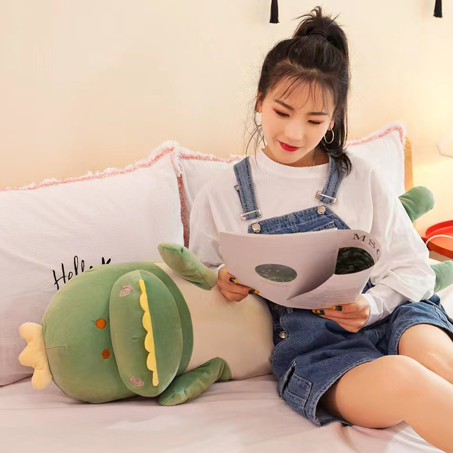 Una giovane donna in salopette di jeans legge un libro seduta su un letto accanto a un grande dinosauro di peluche verde.
