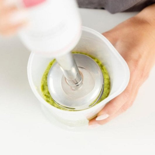 Un frullatore a immersione bianco frulla cibo verde in un contenitore bianco.