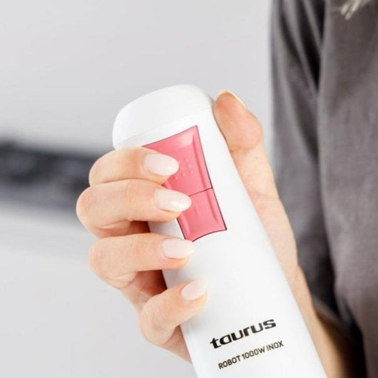 Una mano tiene un frullatore a immersione bianco con pulsanti rosa e marchio 'Taurus'.