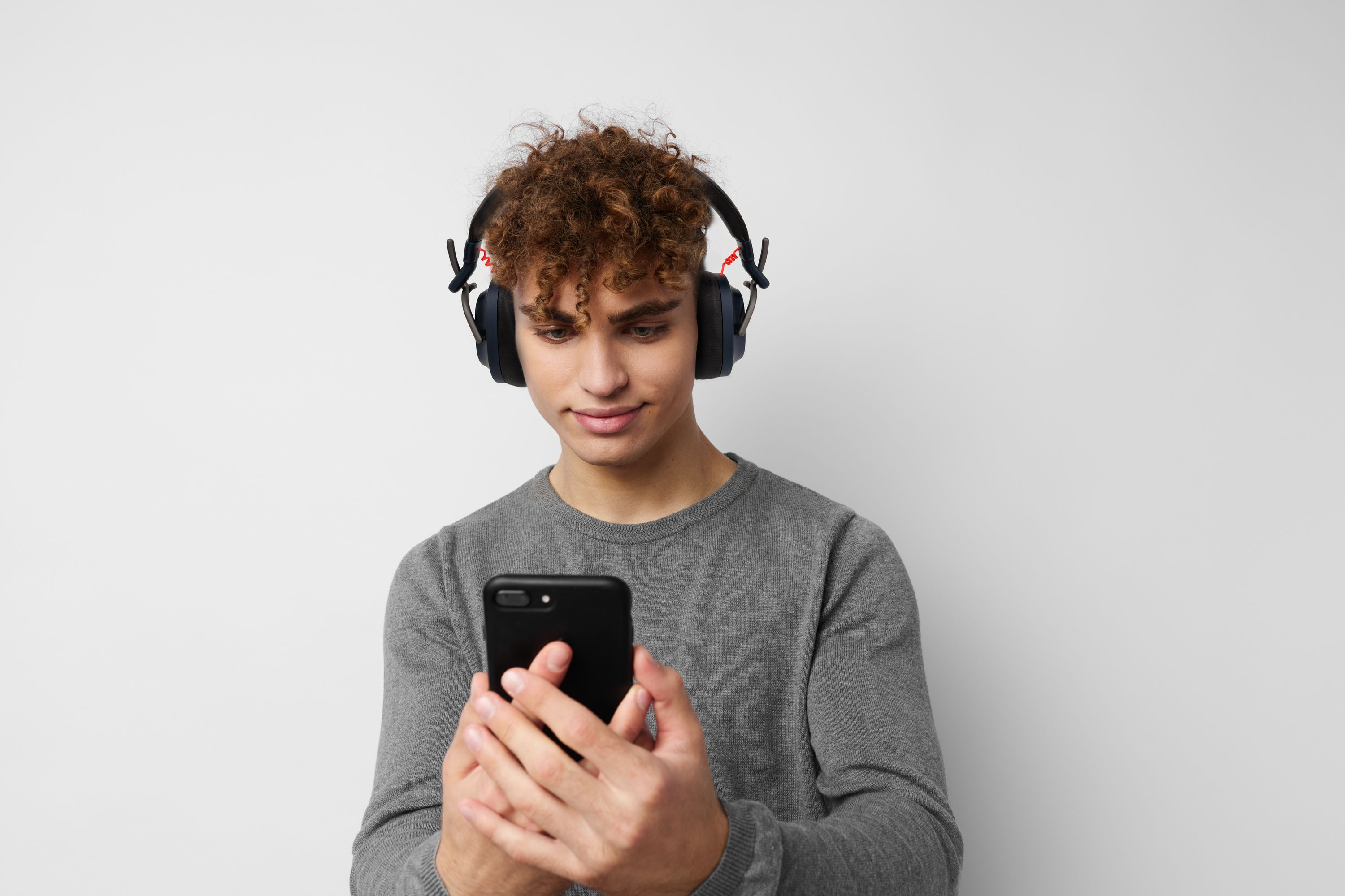 Ein junger Mann mit lockigen Haaren trägt Kopfhörer und schaut auf sein Smartphone.