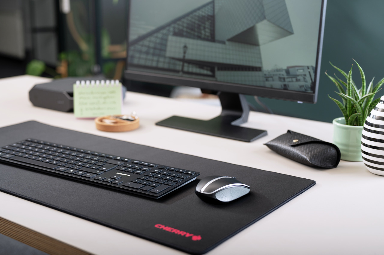 Eine schwarze kabellose Tastatur und Maus auf einer Schreibtischunterlage, mit Monitor und Pflanze.