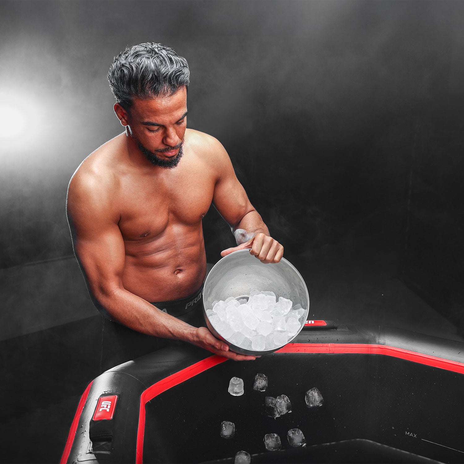 Un hombre con pelo oscuro y barba vierte cubitos de hielo de un bol en una bañera negra.