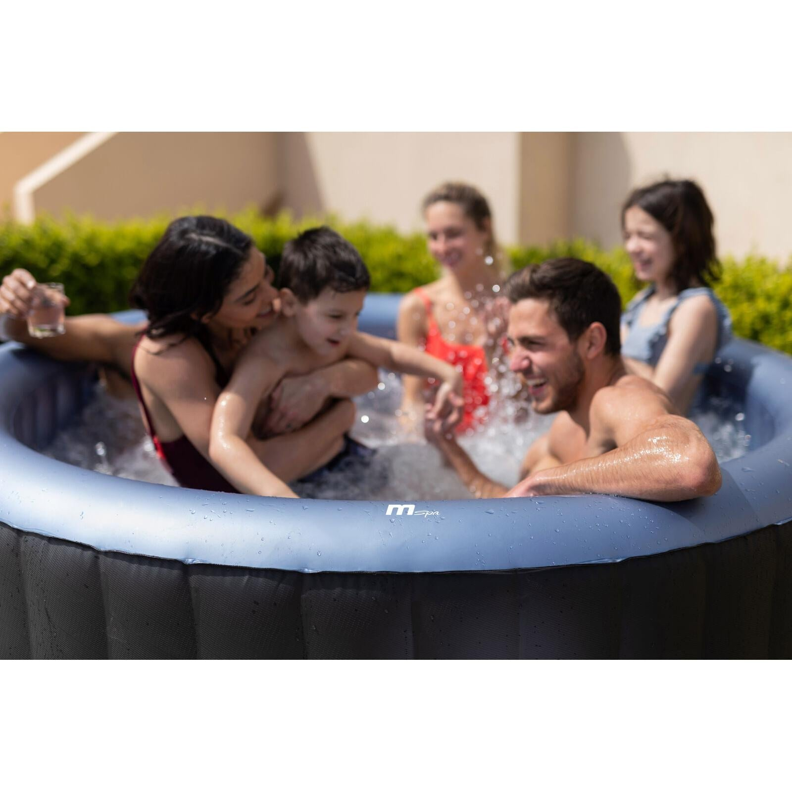 Una familia salpica en un jacuzzi inflable con agua burbujeante y arbustos verdes al fondo.