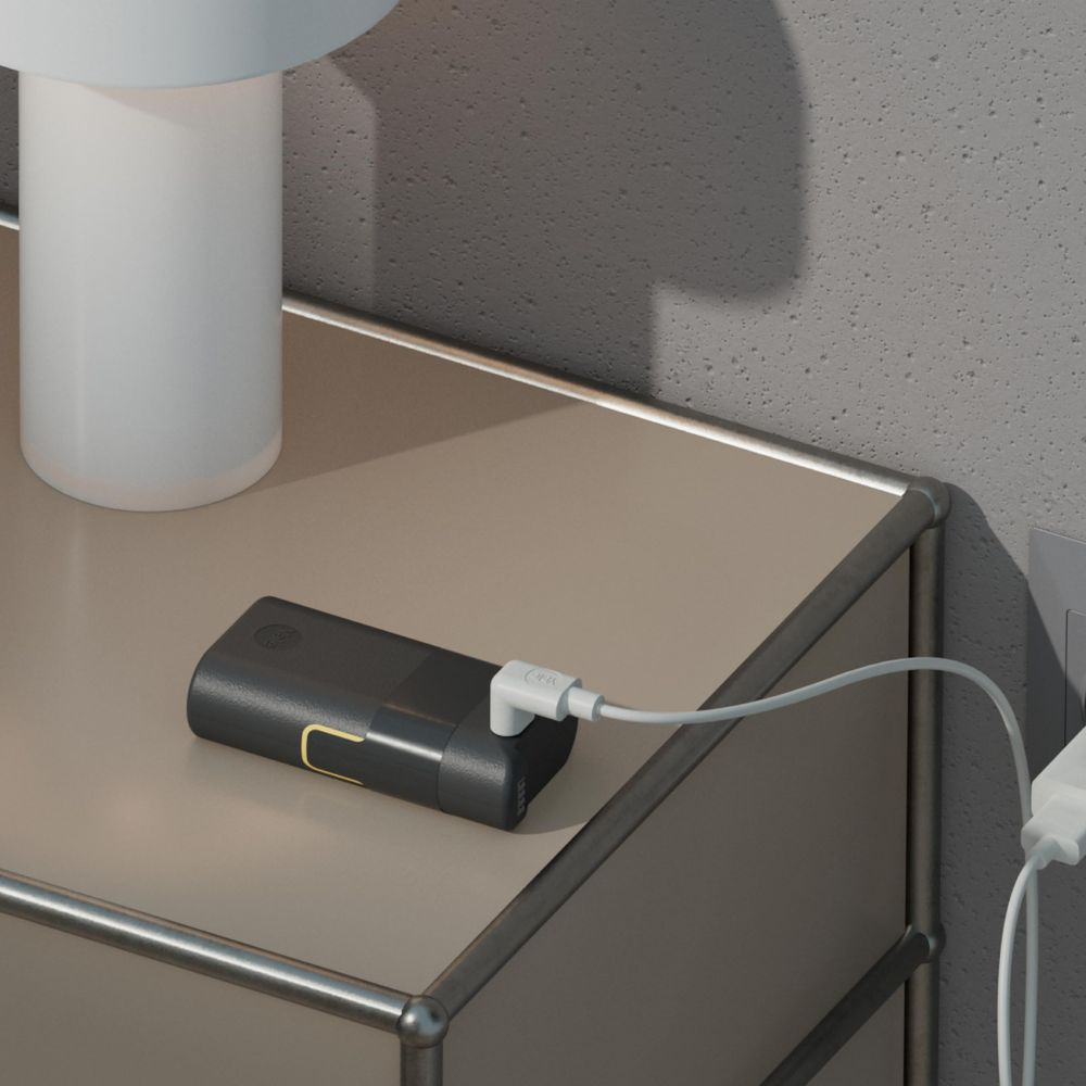 Ciemny powerbank z białym kablem podłączonym do gniazdka, leżący na stole.
