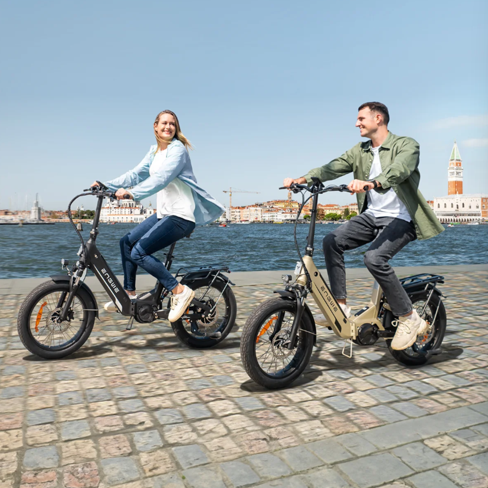 Ein Mann und eine Frau fahren E-Bikes am Wasser entlang mit Gebäuden im Hintergrund.