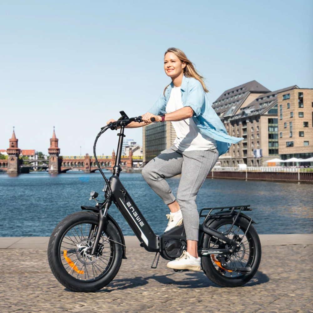 Eine Frau fährt ein schwarzes E-Bike am Wasser mit Gebäuden und Brücke im Hintergrund.