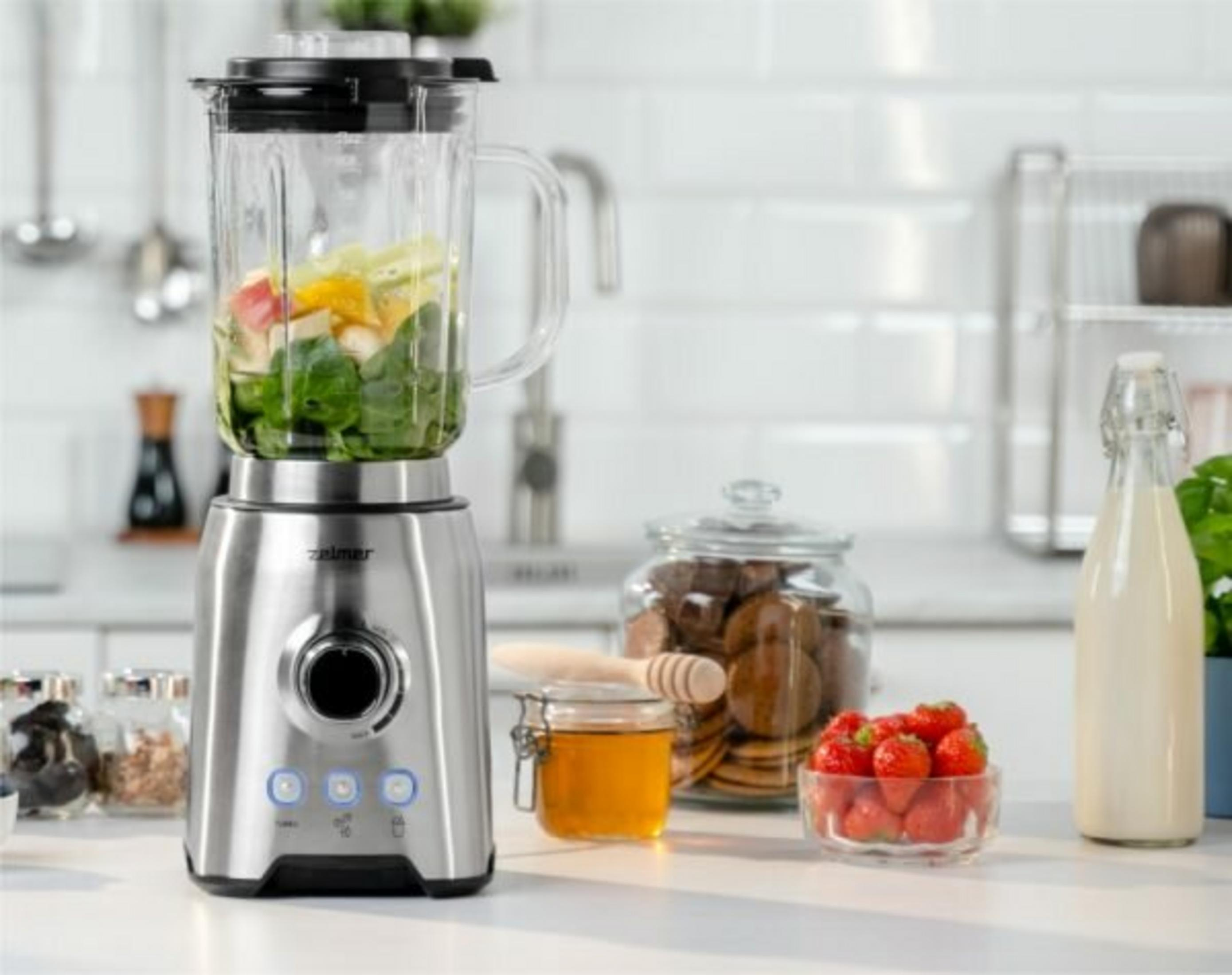 Srebrny blender z owocami i szpinakiem, obok miodu, truskawek i ciastek.