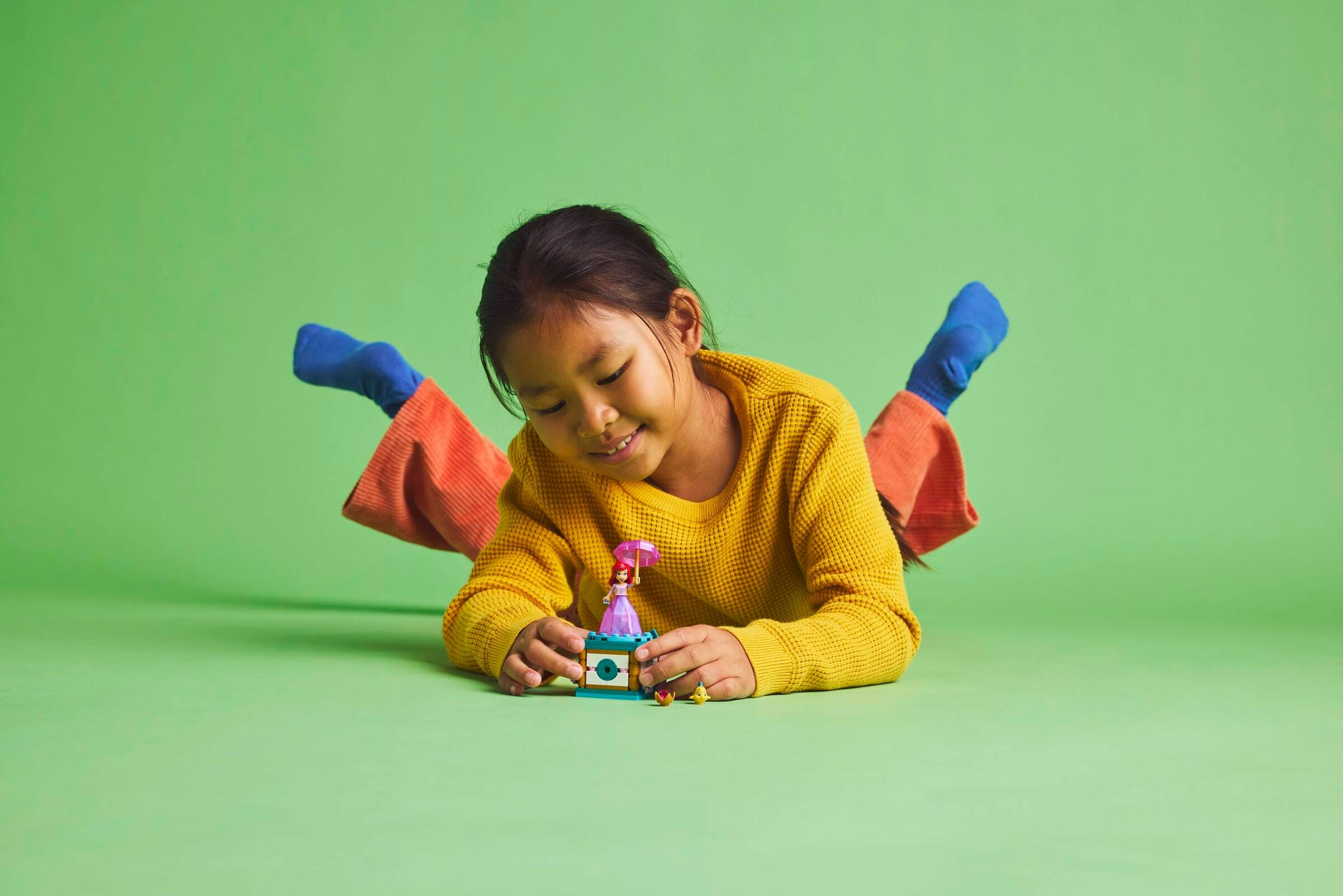Ein Mädchen im gelben Pullover und orangefarbener Hose spielt mit einem Lego-Set auf grünem Hintergrund.