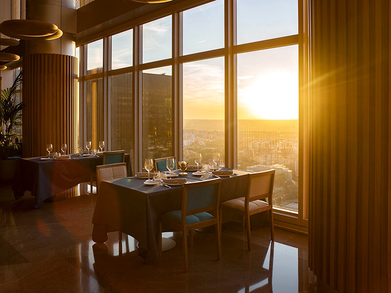 Mesas de comedor de restaurante puestas para cenar con vista al atardecer sobre un paisaje urbano.